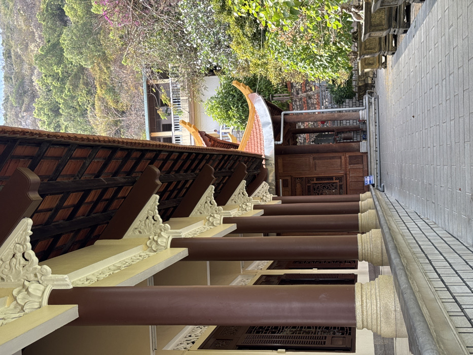 The image displays a long, covered outdoor walkway or veranda, seen from a slightly elevated perspective, looking forward along its length. The photo appears to have been taken in portrait mode and is rotated 90 degrees clockwise.
In the immediate foreground, to the left (which would be the top if rotated correctly), is the underside of a traditional tiled roof or overhang. Dark wooden beams form a grid pattern, supporting reddish-brown roof tiles. Beneath this roof structure, a series of light-colored, decorative corbels or brackets support the eaves. These corbels feature intricate, carved floral or organic motifs in white or very light yellow.
Below these decorative brackets, a series of thick, round pillars run parallel to the view, extending into the distance. These pillars are a dark reddish-brown color, likely wood or painted to resemble wood. They are evenly spaced, creating a rhythmic repetition. The base of each pillar, where it meets the ground level of the walkway, is also light-colored and appears to have some decorative carving.
The walkway or floor itself, running alongside the pillars, is made of light grey square tiles, laid in a grid pattern. To the right of the pillars, further into the scene, a slightly darker, checkered patterned surface is visible, possibly another type of paving or a drain.
In the mid-ground and background, behind the pillars and walkway, a lush natural landscape with dense green trees and foliage is visible. Some parts of the trees show lighter green leaves, suggesting new growth, and a few hints of purple or pink flowers can be seen among the greenery. Beyond the trees, what appears to be part of another building or structure with a lighter-colored wall is visible.
Towards the right side of the image, closer to the background, there's another section of a building with a curved, upturned roof typical of traditional Asian architecture. This section also features dark wooden elements and intricate carvings on what might be a doorway or window frame. A system of pipes, possibly for drainage or utilities, runs along the wall of this building. There appears to be a small blue sign with white text on it near the pipes, but the text is not legible.
The lighting suggests it is daytime, with bright sunlight creating some highlights and shadows, particularly on the roof tiles and the foliage. The overall impression is one of a serene, possibly temple or traditional garden setting, characterized by detailed craftsmanship and natural beauty.
