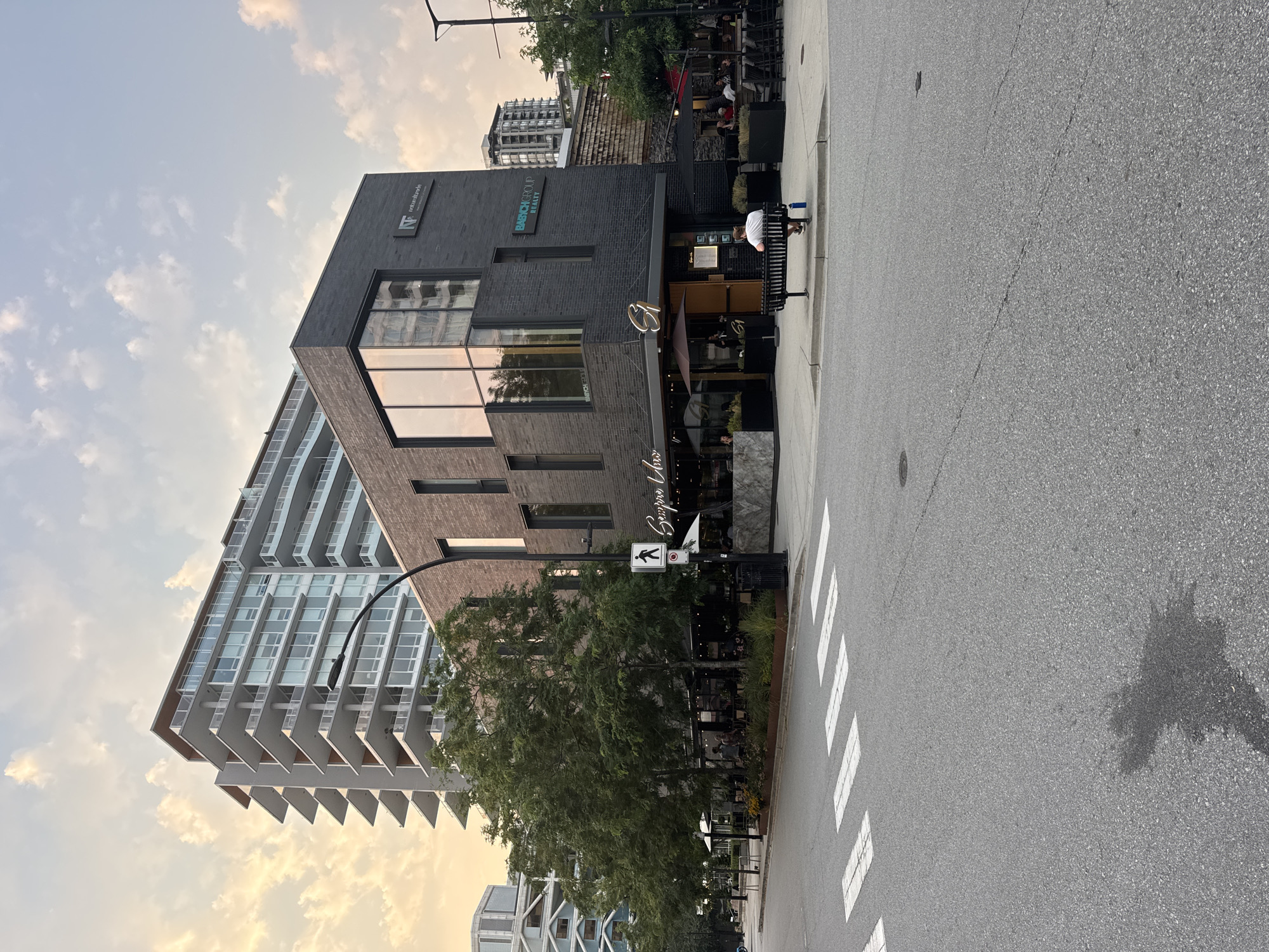 The image shows a street view in North Vancouver, Canada, with a contemporary brick building in the foreground housing a restaurant called "Semprè L'Oro" and a real estate group. A lone person is seated on a bench in front of the building. Behind this building, a modern high-rise apartment complex with multiple balconies is visible. The sky exhibits hues of sunset, with clouds scattered across it. The scene also includes a crosswalk, trees, outdoor seating for the restaurant, and streetlights.