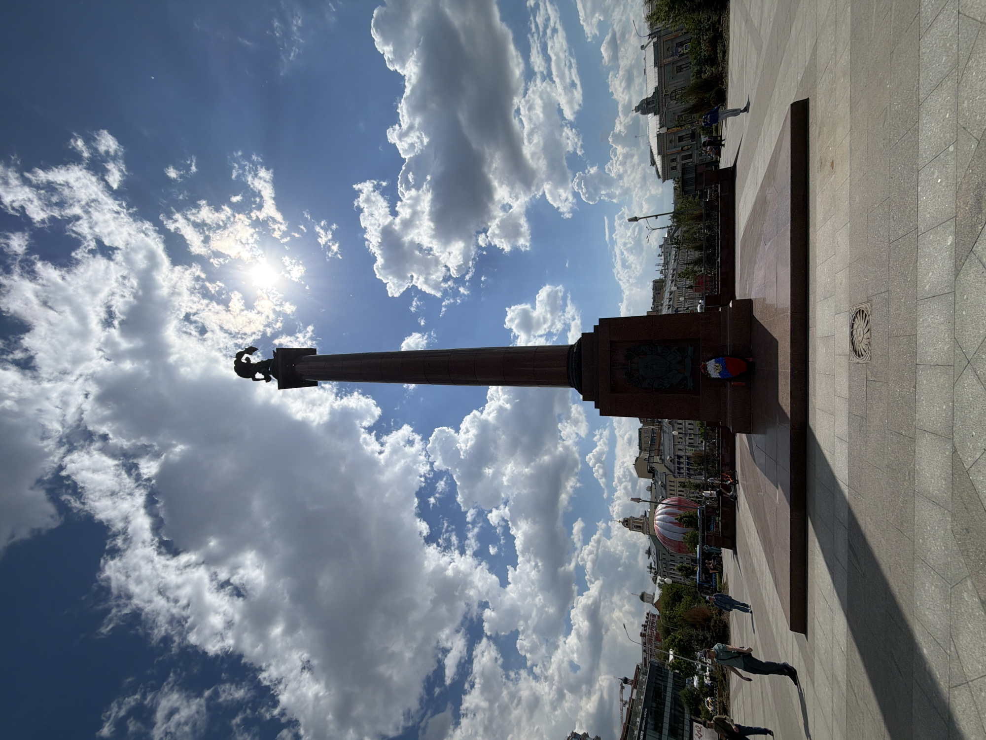 The image captures a vibrant, sunny day in a public square in Moscow, Russia. A tall, reddish-brown stone column, resembling an obelisk, dominates the center of the frame, topped with a dark silhouette of a statue, likely a double-headed eagle, a symbol of Russia. The column rests on a broad, multi-tiered stone base. On one side of the base, a circular wreath adorned with ribbons in red, white, and blue—the colors of the Russian flag—is visible, suggesting a commemorative monument.
The square itself is paved with light grey and reddish-brown tiles. Several people are seen walking across the plaza, their distinct shadows cast on the ground, indicating bright sunshine and a high sun position.
In the background, a cityscape unfolds with various buildings, some with traditional architectural elements. A notable structure features a large, distinctive red and white striped dome or a balloon-like installation on its roof. Other buildings, streetlights, and some trees line the perimeter of the square.
The sky is a brilliant blue, partially covered by scattered, puffy white cumulus clouds. The sun shines intensely, creating a bright glare and lens flare, illuminating the scene. The strong shadows and clear visibility suggest it is midday or early afternoon with fair weather.