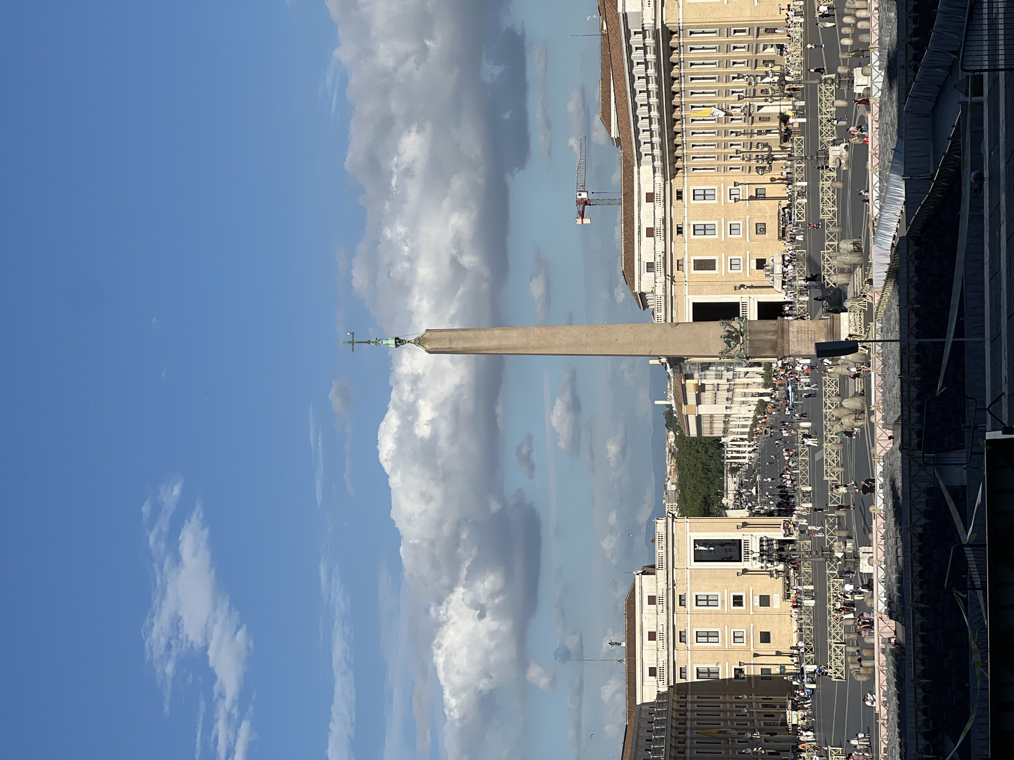 This image depicts a wide view of St. Peter's Square in Città del Vaticano, Italy, under a partly cloudy sky. The central feature is the tall, reddish-beige Egyptian Obelisk, topped with a cross on which a white bird is perched.
In the immediate foreground, a dark, multi-tiered structure, possibly temporary seating or an excavated area, is visible, enclosed by yellow and white tape barriers. A solitary bird stands on the ground level to the bottom right.
The main expanse of St. Peter's Square is visible beyond these foreground structures, filled with numerous pedestrians walking in various directions. The square features light-colored paving, flanked by decorative lampposts and numerous metal crowd barriers and stone bollards.
On both sides of the square, large, multi-story buildings with classical architecture are prominent. The building on the left exhibits multiple windows and a large dark banner or screen on its facade. The building on the right features a repeating series of arches or windows, indicative of the colonnades, and a yellow and white flag (Vatican flag) is visible on a pole.
In the mid-distance, beyond the main square and buildings, further city structures are visible, leading towards a line of distant, hazy hills or mountains. A large red and white construction crane is visible on the horizon to the right of the obelisk. The sky above is a bright blue, interspersed with large, fluffy cumulus clouds that cast some soft shadows on the landscape, suggesting a bright day.