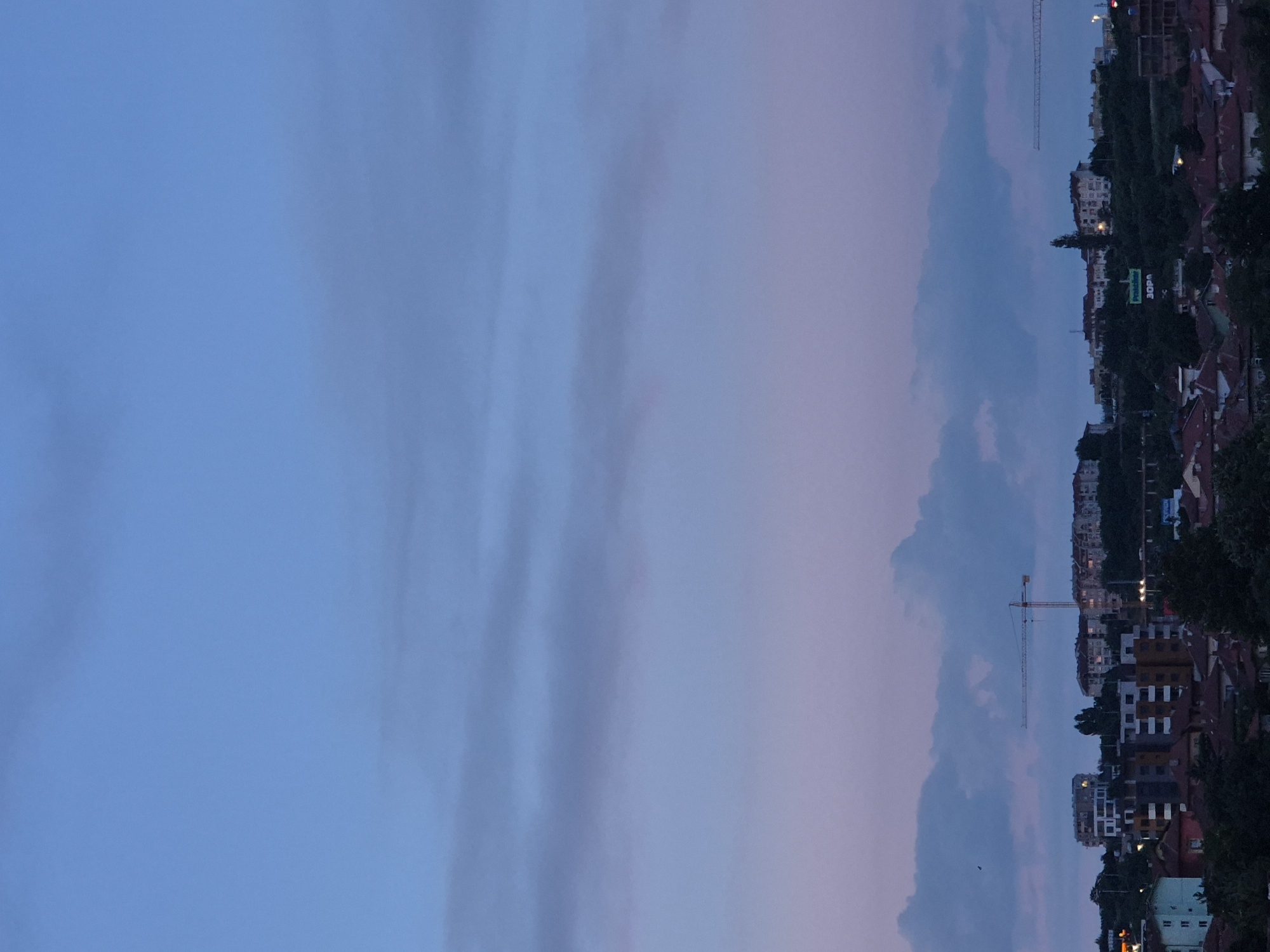 The image depicts a city skyline at dusk. The sky is a gradient of blue and purple, with scattered clouds. In the foreground, there are rooftops of residential buildings, some with red tile roofs. Beyond the rooftops, several multi-story buildings are visible, some appearing to be apartment complexes. A construction crane stands prominently in the middle ground. In the background, more buildings and trees are silhouetted against the twilight sky. A green billboard with white text is also visible on a hill. The overall impression is one of an urban environment at the end of the day.