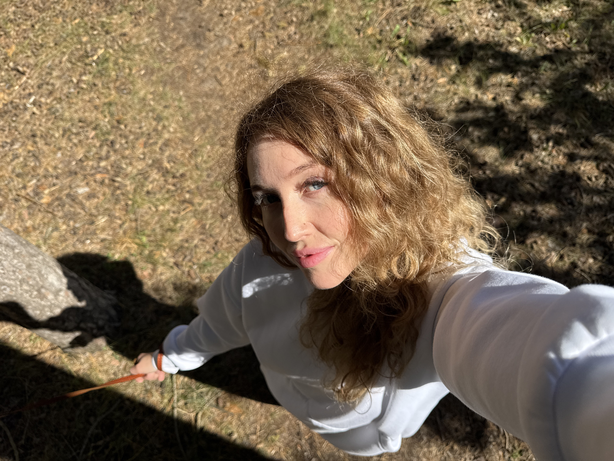 A high-angle medium shot shows a woman from the chest up, looking up and to her right, with her head tilted. She has light skin, blue eyes, and wavy, light brown hair. She is wearing a white sweatshirt. Her right hand is visible, holding a thin, orange leash that extends upwards out of frame. The background consists of dry, brown grass, with a tree trunk and shadows to the upper left and right. The lighting suggests it is daytime, with sunlight casting distinct shadows. The location is not specified, and no elements in the image definitively place it in Chaikovsky, Russia.