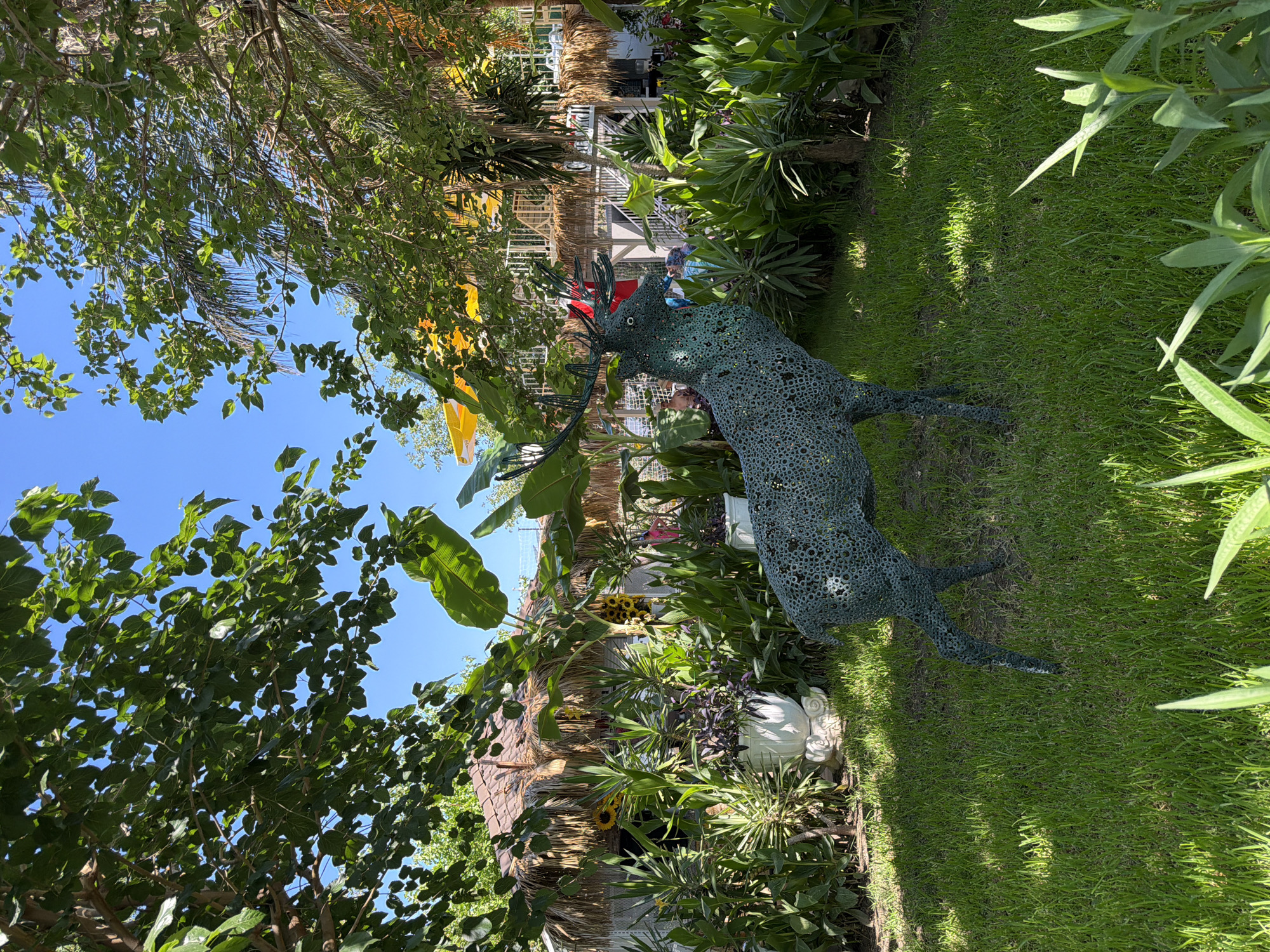 In a lush, green garden area, a dark green, wire-frame sculpture of a deer stands prominently on a manicured lawn. The deer sculpture is detailed, with an openwork design that reveals the surrounding foliage through its body. It has antlers and appears to be facing left. Behind the deer, a thatched-roof structure, possibly a small building or a gazebo, is visible, adorned with sunflowers. Various plants, including large-leafed tropical varieties and spiky succulents, create a dense, verdant backdrop. In the distance, a multi-level structure with white railings and yellow awnings can be seen, suggesting an outdoor seating or entertainment area. The sky above is a clear, bright blue. The scene appears to be set in Kemer, Turkey, judging by the architectural style of the thatched-roof building and the presence of tropical and Mediterranean vegetation common in the region.