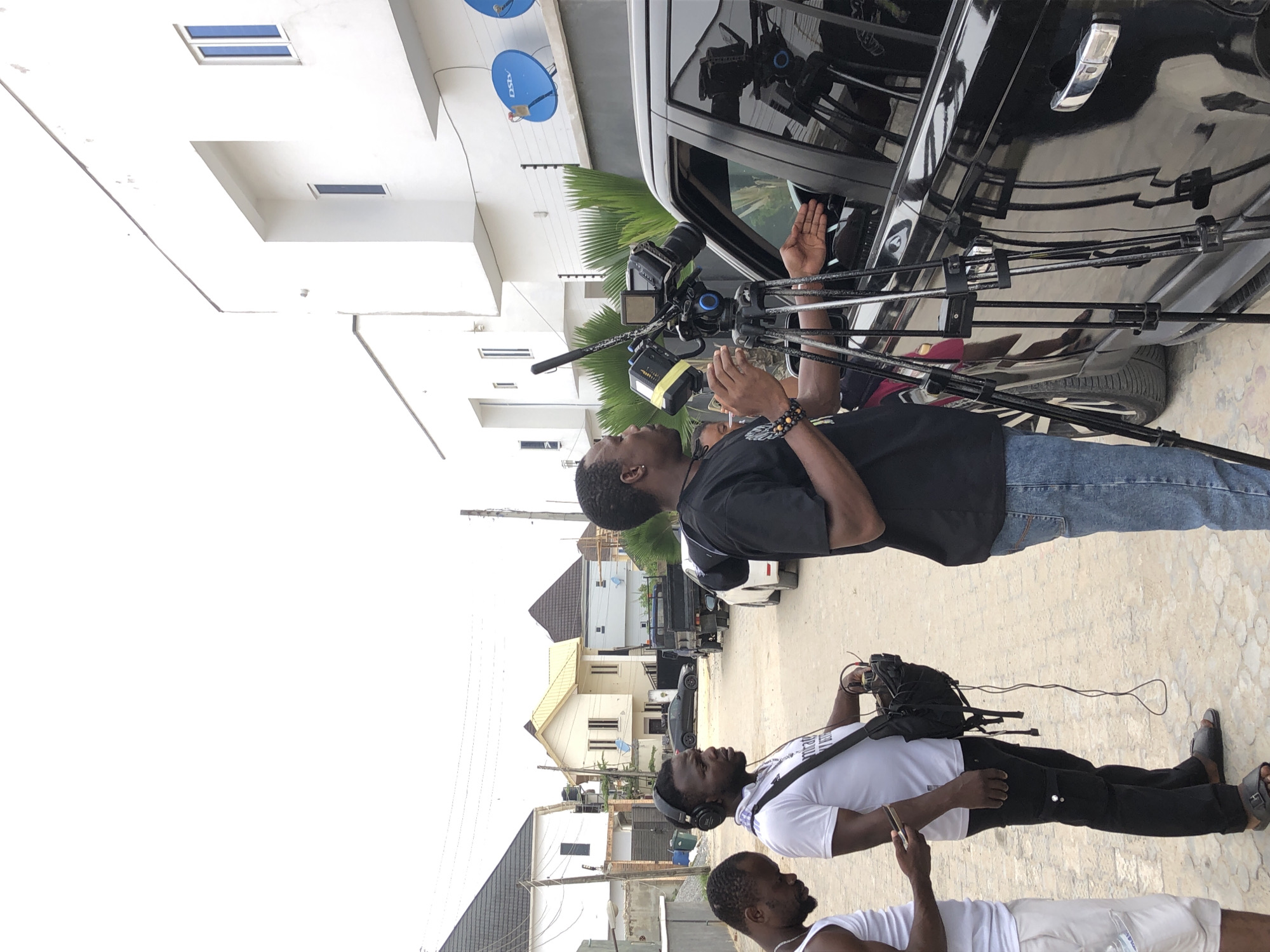 The image, rotated 90 degrees clockwise, captures an outdoor scene, likely a residential street, where a video production appears to be taking place.
In the foreground, three men are present. One man with dark skin, wearing a black T-shirt and blue jeans, is operating a professional video camera mounted on a tripod. His hands are near the camera, which is positioned next to the passenger side of a black SUV or car. He wears a beaded bracelet on his left wrist. The camera is pointed towards the car's window, and another person's hands are visible inside the vehicle, possibly interacting with the camera or being filmed.
Standing to the left of the camera operator is another man with dark skin and a beard, wearing a white T-shirt, black pants, and headphones. He is holding a shoulder bag that likely contains audio equipment, suggesting he is part of the sound crew. Further in the foreground, a third man, also with dark skin and a beard, is partially visible, wearing a white tank top and white shorts, looking at a smartphone.
The background features several modern-looking residential buildings with light-colored walls and dark roofs. On one of the white buildings, two blue satellite dishes with "DStv" branding are clearly visible. Palm trees are also seen, adding to the tropical or subtropical setting. The sky is bright, indicating it is daytime. The ground is a paved or concrete surface. The overall impression is a focused film or video shoot in progress.