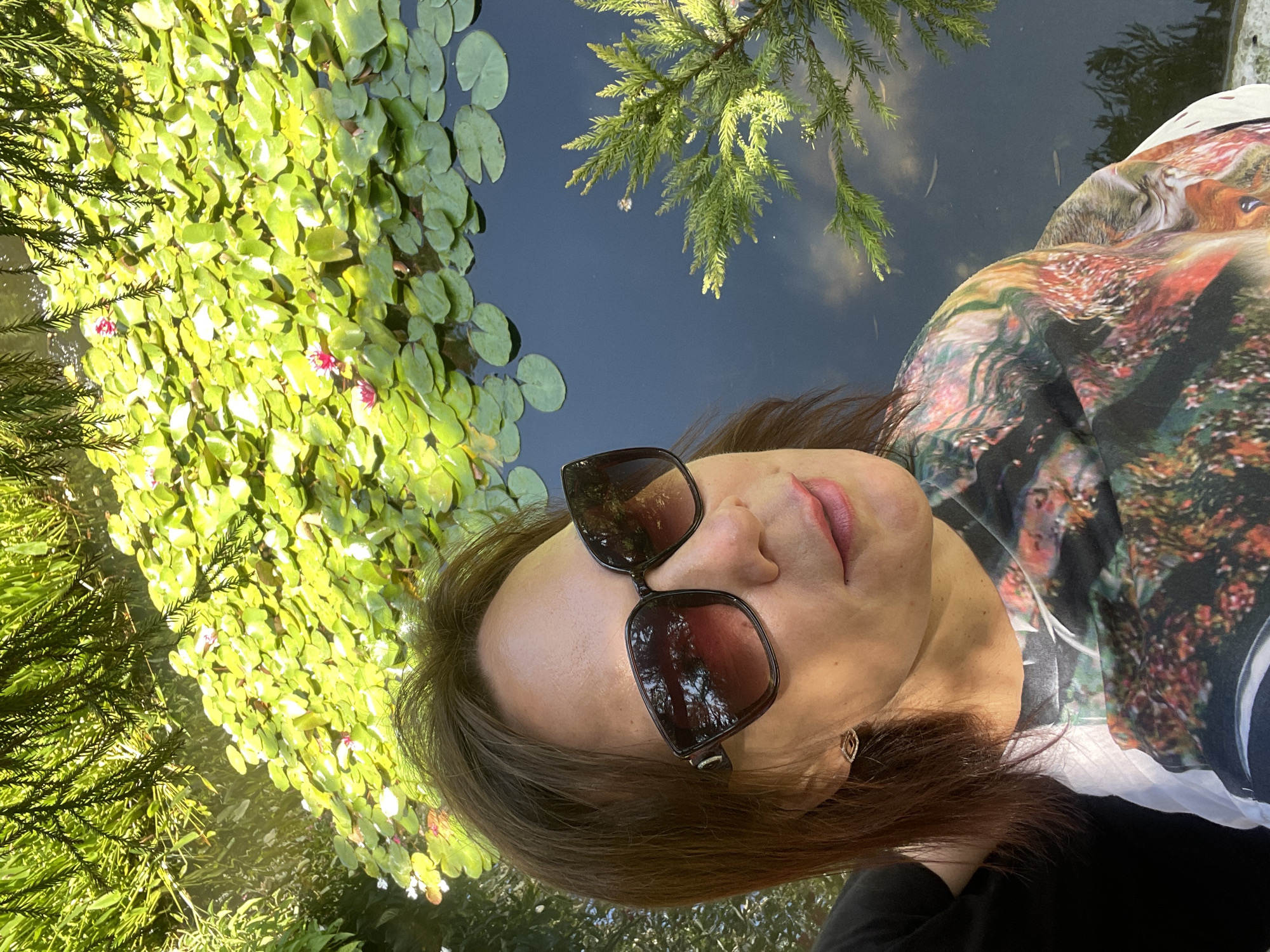 A woman wearing large, dark sunglasses and a colorful shirt stands in front of a pond filled with lily pads. The pond is dark and reflective, with some trees and foliage visible in the water's reflection. There are pink water lilies blooming on some of the pads. The woman has medium-length brown hair and is wearing a pair of small, gold earrings. The image appears to have been taken outdoors on a sunny day. The overall scene suggests a peaceful, natural setting.