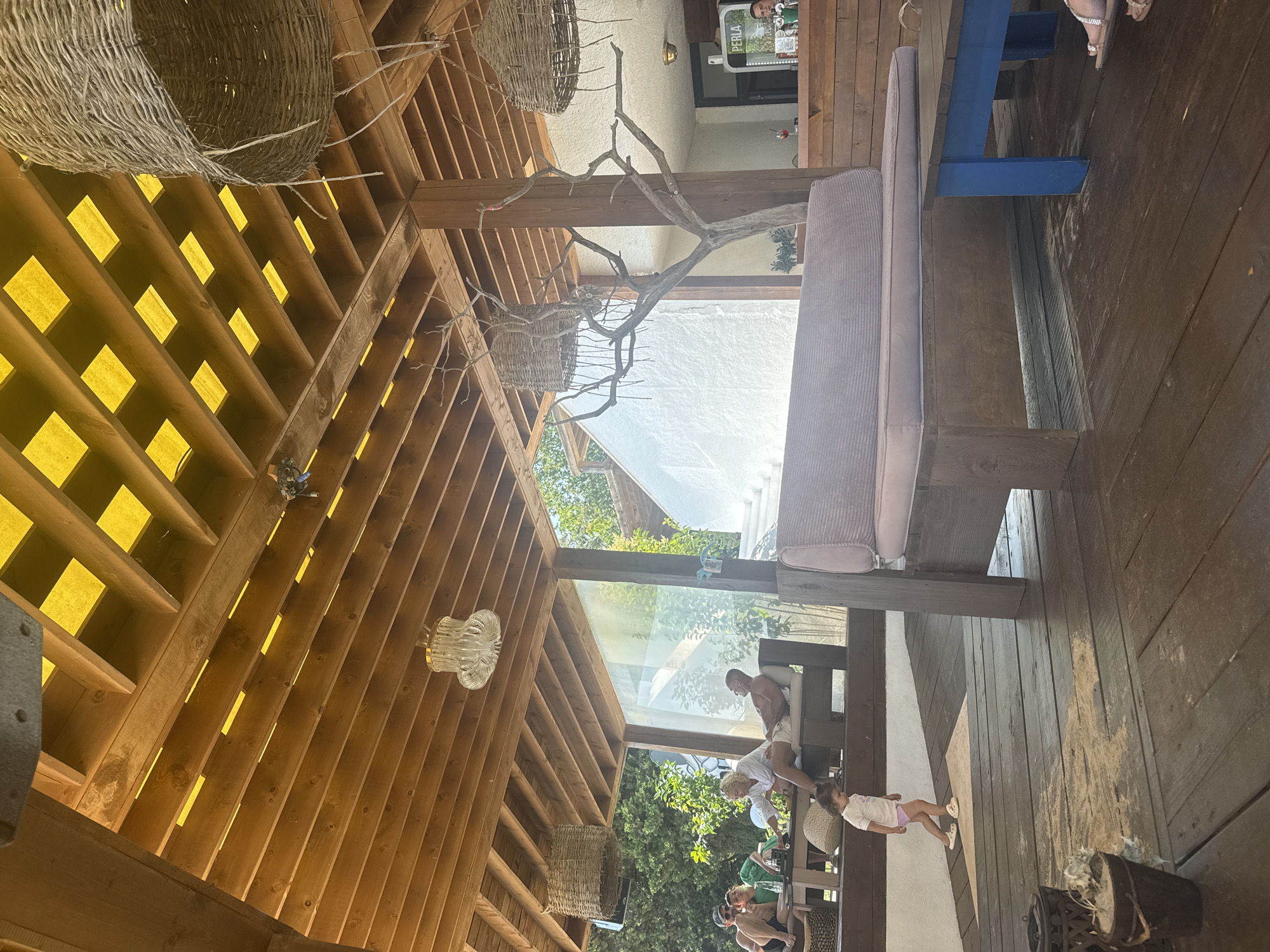 A low-angle, sunlit outdoor scene depicts a rustic wooden veranda at a establishment in Primorsko, Bulgaria, with a visible yellow ceiling structure above. In the foreground, a young child in a white t-shirt and purple shorts walks across a weathered wooden floor. To the right, a substantial wooden bench with a light purple upholstered seat faces a blue-legged wooden table. Further back, on the left, a group of adults are seated on loungers and at a table, appearing to be socializing. A glass partition separates this seating area from a white-washed structure with visible stairs. The background reveals lush green trees. Decorative woven hanging lamps and a bare tree branch are suspended from the wooden ceiling. A cooler labeled "PERLA" and a bar area are visible in the far background, with a bartender present. The overall ambiance is relaxed and summery.