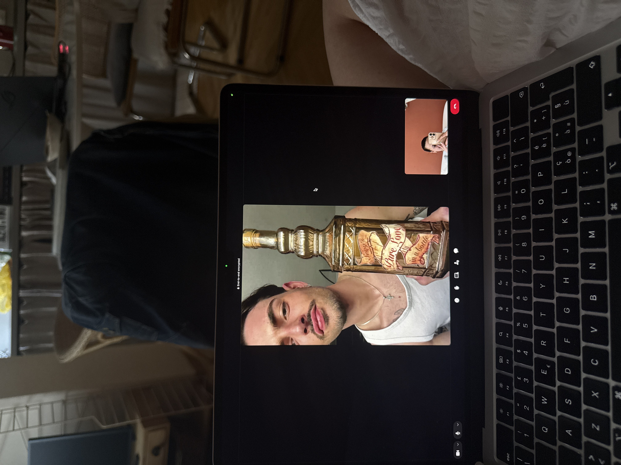 Here is a forensic description of the image:
A person is having a video call on a laptop. The laptop's keyboard is visible. On the screen, a man is holding up a bottle labeled "Pure Love in a bottle". The other person in the video call is visible in a smaller window in the upper right corner of the screen. The location appears to be indoors, with a bed or couch and a chair visible in the background.