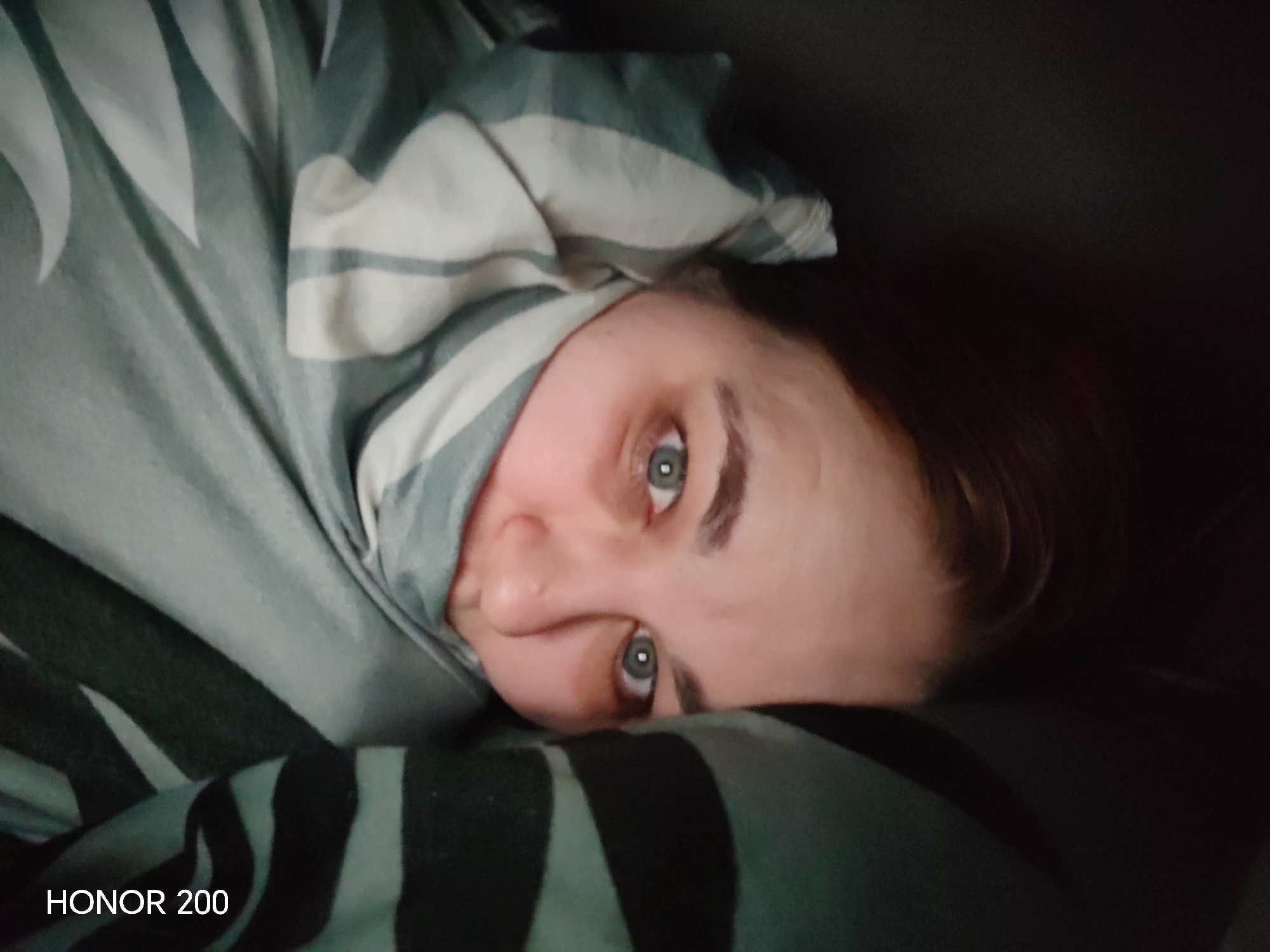 An indoor photograph, captured in the city of Chaikovsky, Russia, depicts a single individual, appearing to be an adult female, in a close-up, horizontal orientation. The subject's head and upper torso are visible, with her face turned directly towards the camera. She possesses light skin, dark eyebrows, and light-colored eyes (consistent with blue or green irises) with visible pupils. A portion of dark hair is discernible on the right side of her head. Her expression is neutral.
The individual is largely enveloped in bedding, specifically a blanket or duvet. This bedding features a pattern of light green/teal, white, and darker green/black stripes and abstract shapes. One section of the bedding is wrapped around her head and upper body, while another lies beneath her chin and neck. The background to the right of the subject is dark and indistinct. The text "HONOR 200" is digitally overlaid in white font at the bottom left corner of the image. The scene suggests an indoor environment, likely a bedroom, with low or indirect lighting.