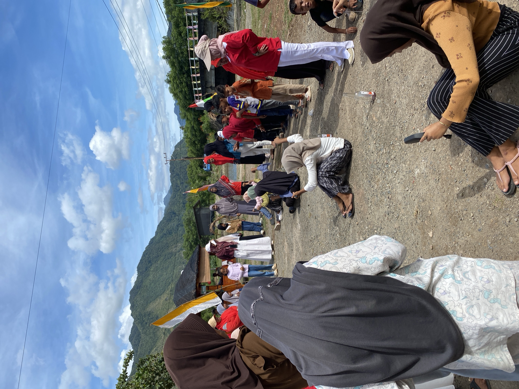 In Aneuk Paya, Indonesia, a group of people, primarily women and children, are gathered outdoors on a gravelly surface. The scene appears to be a casual event or gathering, possibly a competition, given the presence of several flags and people seated on the ground.
In the foreground, a woman wearing a brown hijab and an orange long-sleeved shirt with striped black and white pants is seated on the ground, looking at a black object in her hand, possibly a phone or a small mirror. To her left, another woman in a dark grey hijab and a white shirt with a unicorn pattern is also seated, facing away from the camera.
A line of children and adults, mostly in red and white attire, are standing in the middle ground, facing a different direction. Some children are participating in an activity, with one young girl in a light purple shirt and blue pants kneeling on the ground. A woman in a red shirt and white pants stands prominently among the group.
The environment includes a road in the background, a bridge, and lush green hills and mountains under a bright blue sky with scattered white clouds. Several flags, including Indonesian flags, are visible, suggesting a patriotic or celebratory occasion. The overall atmosphere seems relaxed and social.