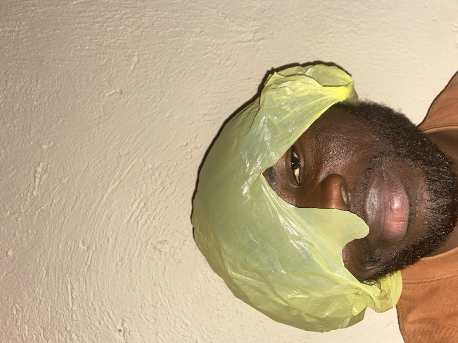 An adult male with dark skin and a beard is depicted in a medium close-up, centered against a light beige, textured wall in Udu, Nigeria. He is wearing a translucent, crinkled yellow plastic bag on his head, which drapes over his forehead and the left side of his face, partially obscuring his left eye and the bridge of his nose. His right eye is visible and directed towards the camera. His mouth is closed. He is wearing a brown or terracotta-colored garment, visible at the bottom of the frame. The lighting source is not directly visible, but illuminates the subject and wall, revealing surface textures.