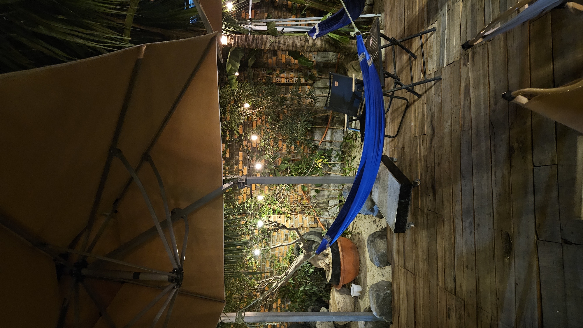 This media file captures an outdoor patio or garden area at night, likely in Cam Đức, Vietnam. The scene features a cozy and relaxed atmosphere, illuminated by warm string lights.
In the foreground on the left, a large, beige patio umbrella extends, with its fabric and support structure visible from below. To the right of the umbrella, a bright blue fabric hammock is suspended, appearing ready for use. Adjacent to the hammock is a dark, foldable outdoor chair.
The background reveals a rustic brick wall partially covered by lush green foliage, including various plants and what appears to be palm fronds. The string lights are integrated into the greenery, casting a soft glow. On the ground, there is a wooden deck floor, and below the hammock, a large terracotta pot holds a sculptural plant or bonsai-like tree, surrounded by several rocks.
The image conveys a tranquil evening setting, perfect for relaxation. No people are visible, and there is no discernible text in the frame.