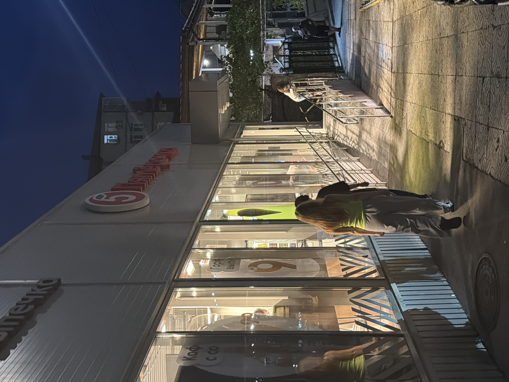 The image depicts an exterior view of a "Pyaterochka" grocery store in Sochi, Russia, at dusk or night. A bright white light beams from the upper right, creating a diagonal line across the dark blue sky and casting shadows on the building's facade. The store's facade is primarily gray with prominent glass windows reflecting the interior lights. A red and white "5" logo is visible on the building.
Inside the windows, several posters are displayed. One poster has Cyrillic text that appears to read "Кофе" (coffee) and "с собой" (to go). Another poster features a large orange "9" and the Cyrillic words "Горяч" (hot) and "выгод" (bargains).
Three individuals are walking away from the camera on a paved walkway adjacent to the store. The person in the foreground is wearing a lime green tank top and gray wide-leg pants. To their left, partially obscured, is another person in darker clothing. Further behind them, a third individual in dark clothing is also walking away.
A ramp with metal railings leads up to an entrance of the store. Further down the walkway, two more individuals are visible. One is a person in a light-colored top and darker pants climbing the ramp, and another is an older man wearing a dark shirt and shorts standing near a metal fence on the right side of the walkway. The ground is paved with textured tiles. Vegetation, including a large leafy vine, grows over a section of the fence on the right. A building with lit windows is visible behind the store in the background.