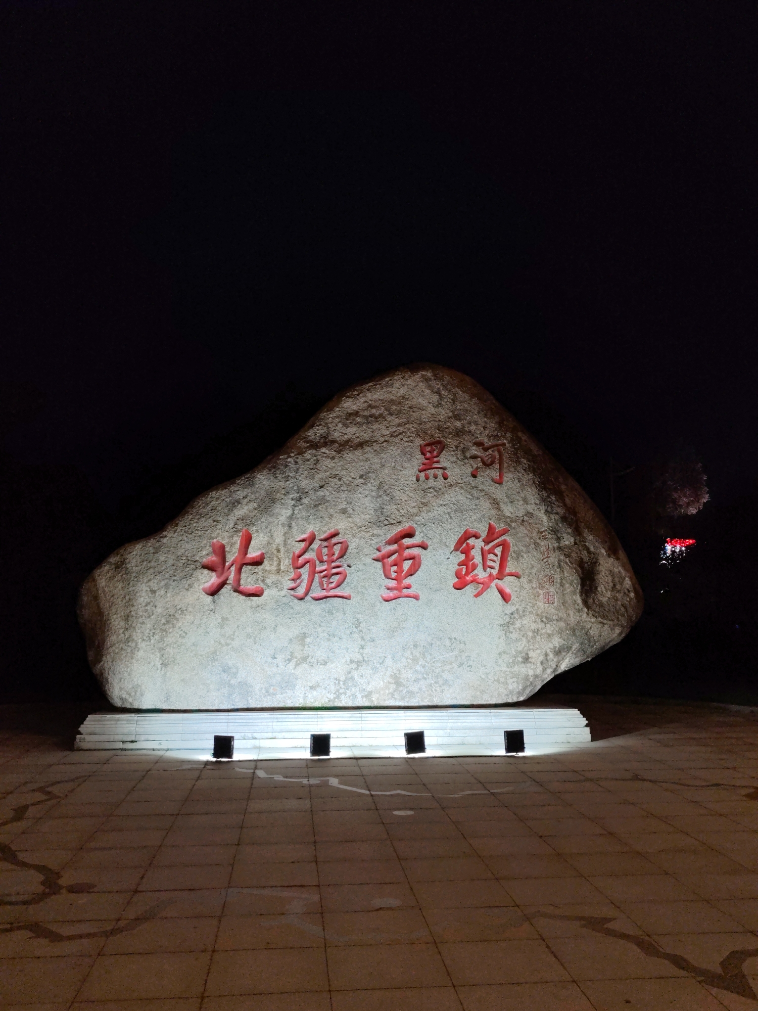The image captures a large, rough-hewn rock monument at night, illuminated by artificial lights. The rock is situated on a paved plaza in 幸福乡, China. The monument is inscribed with red Chinese characters. The top line of characters reads "黑河" (Hēihé), and the larger characters below read "北疆重镇" (Běijiāng zhòngzhèn). The rock is supported by a white, tiered base that has two dark rectangular openings on its front. In the background, to the right of the monument, a faint display of red and white lights suggests fireworks or a distant light show. The plaza pavement is a grid of square tiles, with some water or dark stains creating irregular patterns. The sky is dark, indicating nighttime.