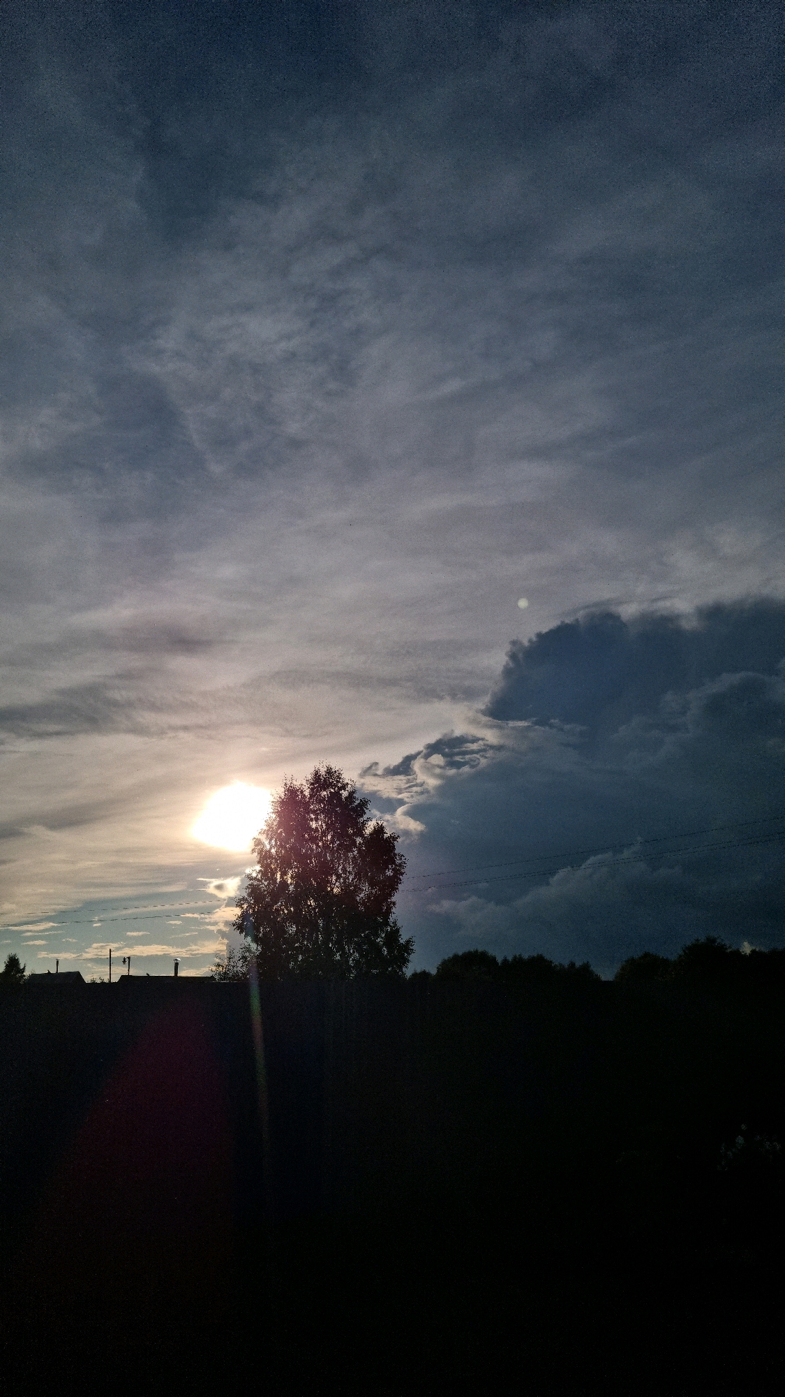 This is a vertical, eye-level, medium shot of a treeline against a dramatic sky over Vysokoe, Russia. The sky is a dominant element, featuring a mix of dark, billowing storm clouds on the right and lighter, wispy clouds interspersed with the bright, setting sun on the left. The sun is a prominent, intense white orb peeking through the clouds, casting a warm glow and creating lens flare streaks. A solitary, medium-sized deciduous tree stands silhouetted against the sun, its branches reaching upwards. Below the treeline, the foreground is entirely black, obscuring any details of the ground or any potential objects or individuals. The silhouette of some buildings with chimneys are faintly visible on the far left, near the horizon. Power lines are visible against the sky.