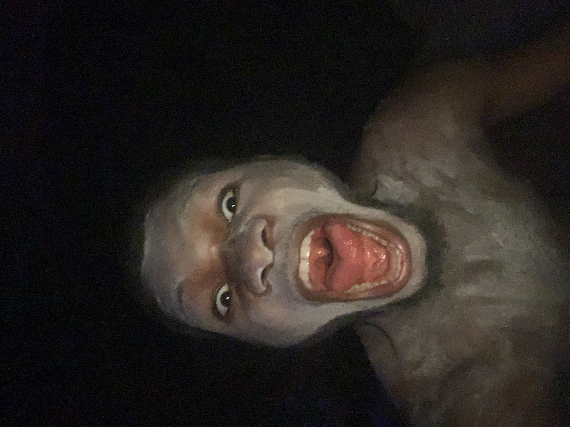 A low-angle photograph features a dark-skinned person with an open mouth, revealing teeth and tongue. The background is black. The subject's face is well-lit, with details visible in the eyes, nose, and skin. The person's upper body is partially visible. No location can be ascertained from the image.