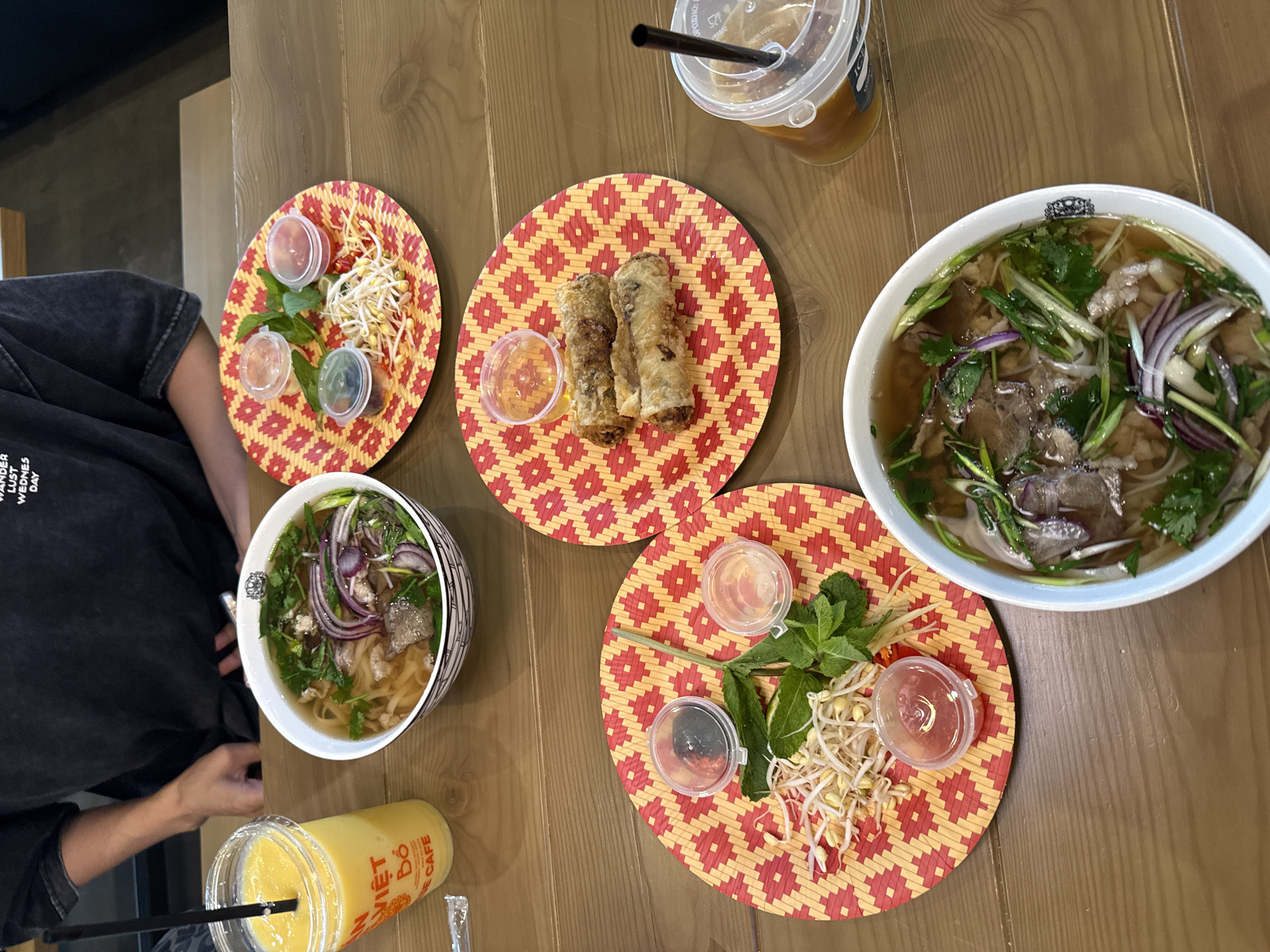 The image presents an overhead view of a Vietnamese meal served on a wooden table at Viet Do Cafe in Krasnoyarsk, Russia. A person, clad in a dark t-shirt with partial white text visible on the chest, is seated in the upper left portion of the frame. The table is set with two large white bowls of pho, each filled with beef, rice noodles, broth, sliced red onions, green onions, and fresh cilantro.
Between the two pho bowls, a round, red and yellow patterned placemat holds two fried spring rolls alongside a small clear container of dipping sauce. Two additional identical placemats are placed near each pho bowl, functioning as garnish plates. Each garnish plate contains a mound of bean sprouts, fresh mint leaves, and a slice of lime, accompanied by three small plastic containers with lids holding various condiments: one red chili sauce, one dark paste, and one clear liquid.
Two drinks are also visible: a clear plastic cup filled with a bright yellow mango smoothie and a black straw, prominently featuring the "VIỆT ĐÓ CAFE" logo; and another clear plastic cup containing an iced brown beverage with a black straw. The wooden tabletop displays a visible grain pattern.