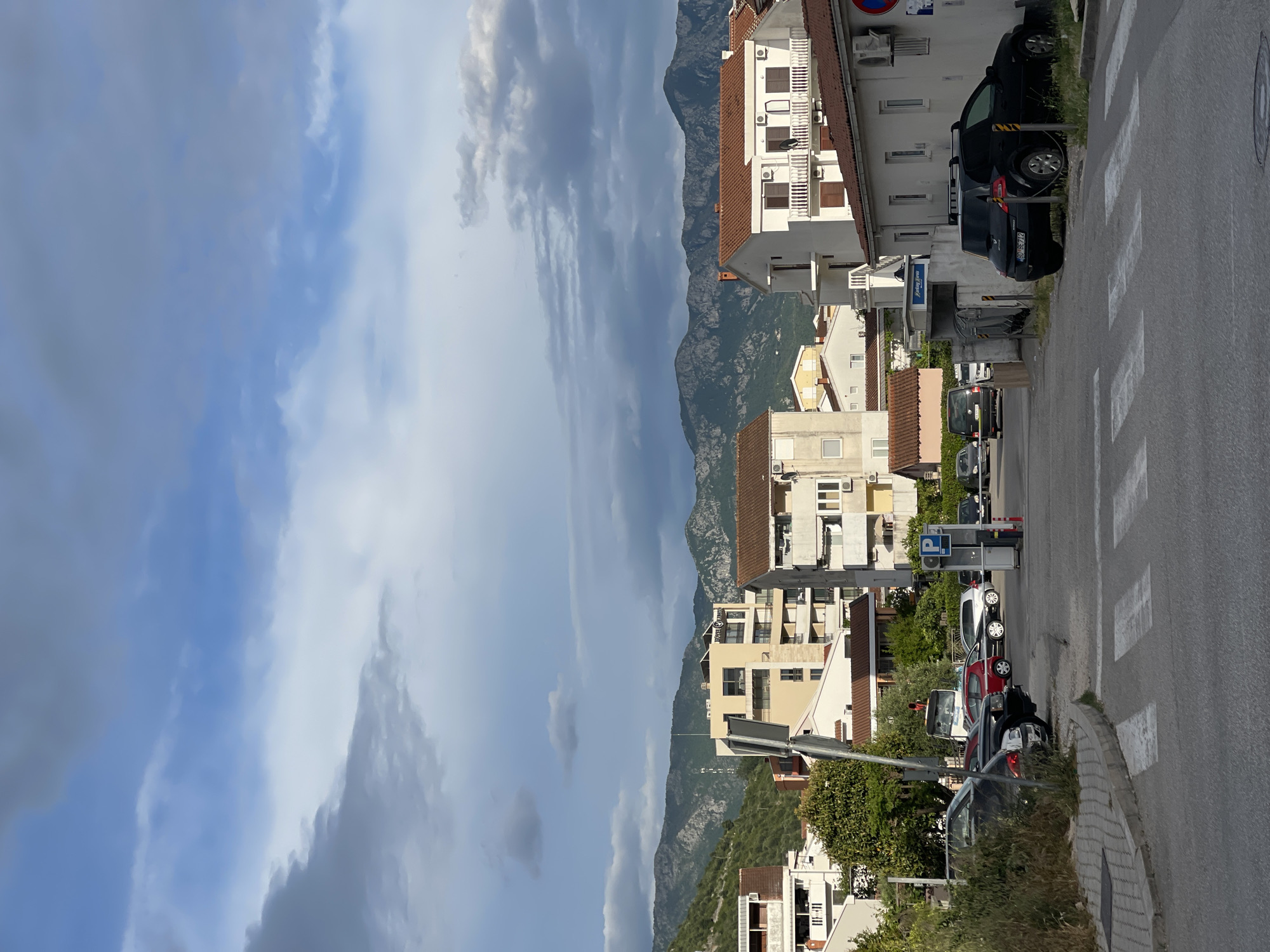 The image shows a street scene in Budva, Montenegro. The photograph is taken from a slightly elevated position looking down a street with parked cars on the right. Buildings line the right side of the street, primarily multi-story residential structures with white or light-colored facades and red-tiled roofs. A mountain range is visible in the background, partially obscured by clouds. The sky is overcast with a mix of blue and gray clouds. A pedestrian crosswalk is visible on the right side of the street. The road is paved, and the weather appears to be daytime.