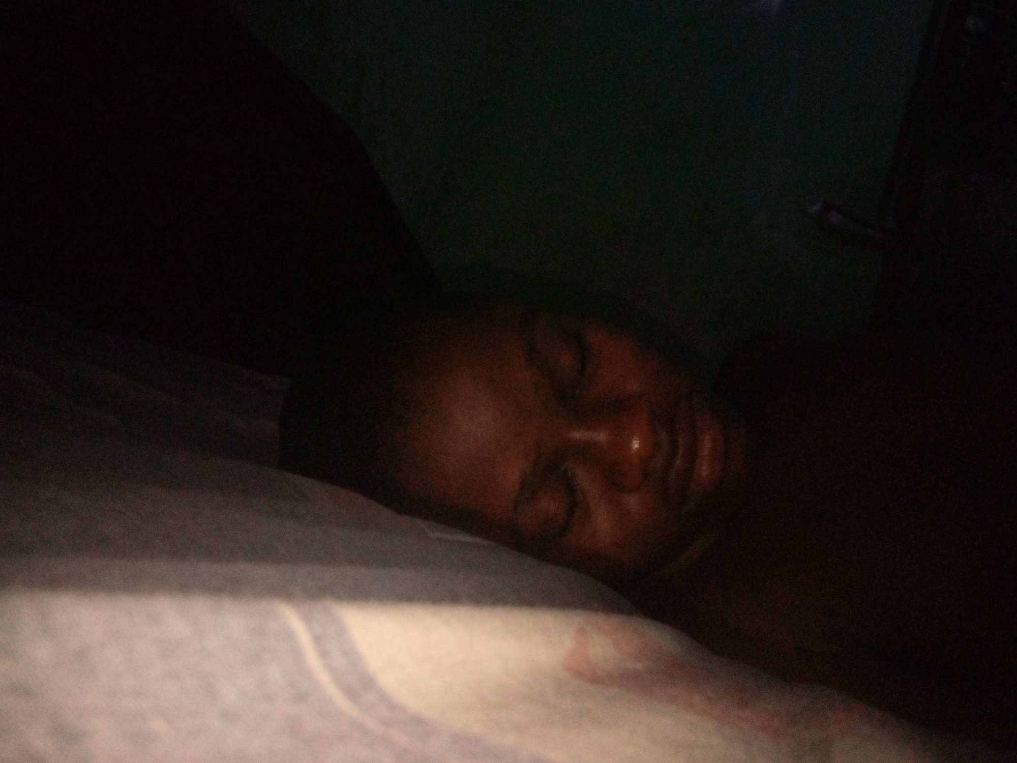 A person's face is partially visible in the foreground, resting on a pillow. The lighting is dim, suggesting an indoor setting, possibly a bedroom. The background is dark and indistinct. The image is composed from a close-up perspective, focusing on the individual's features. The scene appears to be in Shangisha, Nigeria.