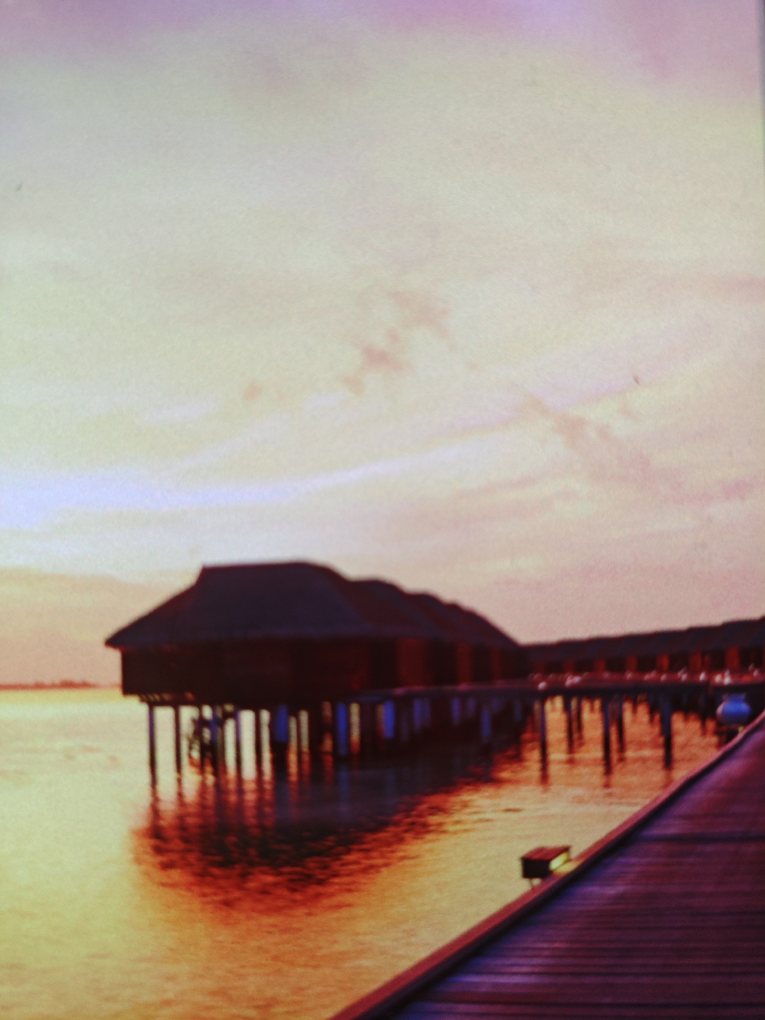 The image depicts an overwater bungalow structure with a wooden walkway extending into the foreground. The scene is illuminated by a warm, sunset-like glow, casting orange and yellow reflections on the water. The sky is a gradient of soft pinks and purples. There are no people visible in the image. The architecture of the bungalow suggests a tropical or resort setting. The image does not provide any specific information or visual cues to associate it with the city of Armavir, Russia, which is geographically distinct from a tropical island environment implied by the subject matter.