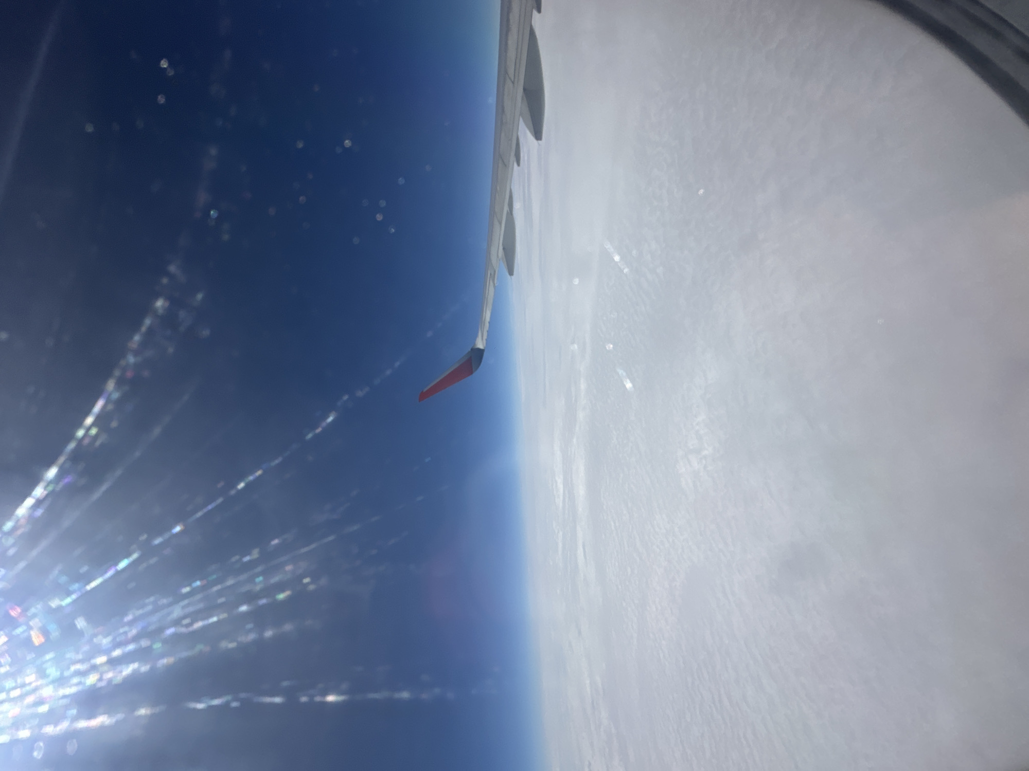 The image shows an aerial view from an airplane window, looking out at the Earth's curvature and the sky. The perspective is rotated, with the airplane's wing prominently featured on the right side of the frame. The wing is a light gray color with visible struts and the tip is painted red. Below the wing, a vast expanse of white clouds blankets the Earth's surface, creating a textured, undulating pattern. To the left of the clouds, the sky transitions into a deep, vibrant blue, indicating a high altitude.
Scattered across the blue sky are numerous bright, bokeh-like orbs of light, as well as streaks of light that appear to be caused by reflections or light refracting through the airplane window. These light effects are particularly concentrated in the lower left portion of the image, suggesting the sun is in that direction and its rays are interacting with the window's surface. There are no discernible people or specific objects other than the airplane wing.
The scene suggests an ongoing flight, likely during daytime, given the bright sunlight and clear blue sky. The thick cloud cover below implies a substantial altitude, obscuring any ground-level details or specific location cues, though the provided context suggests Zhukatau, Russia. The overall impression is one of vastness, altitude, and the visual phenomena associated with air travel.
