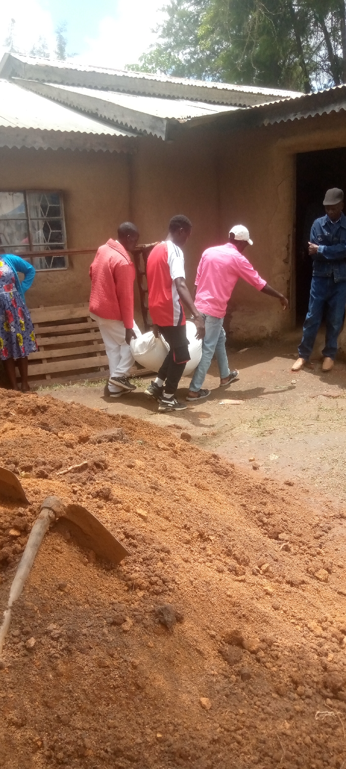 The image shows four men carrying a large white sack in front of a building with a corrugated metal roof, located in Burundu, Kenya, at coordinates 0.46 latitude and 34.776 longitude. The men are wearing casual clothing. One man is wearing a red shirt, another a pink shirt, another a white shirt, and the last man is wearing a red jacket. There is a woman standing in the background, behind the men, wearing a floral dress. The ground is covered in loose, brown dirt and there is a shovel lying on the ground in the bottom left corner of the image. There is a wooden pallet to the left of the men. The scene appears to be taking place in the yard of a residential property. The building is constructed with concrete and has a dirt floor. The scene is bright and the weather is clear.