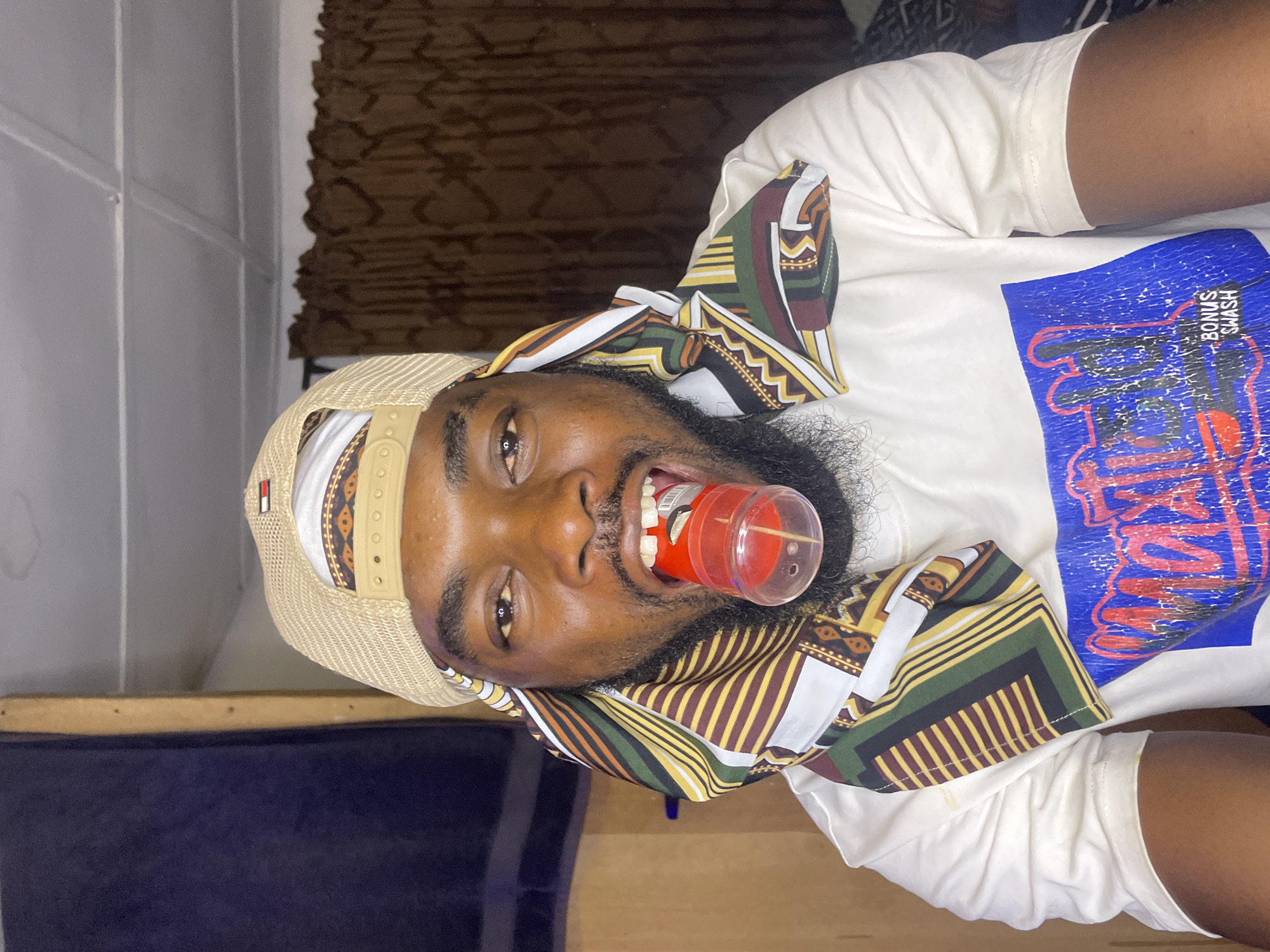 A man is lying down, facing the camera. He is wearing a white t-shirt with a blue graphic print and an African print scarf around his neck. He also wears a tan baseball cap. He has a beard. In his mouth, he is holding a small, red, cylindrical object. The background includes a dark brown curtain and a blue piece of furniture. There is no verifiable location or information that the image was taken in Jos, Nigeria.