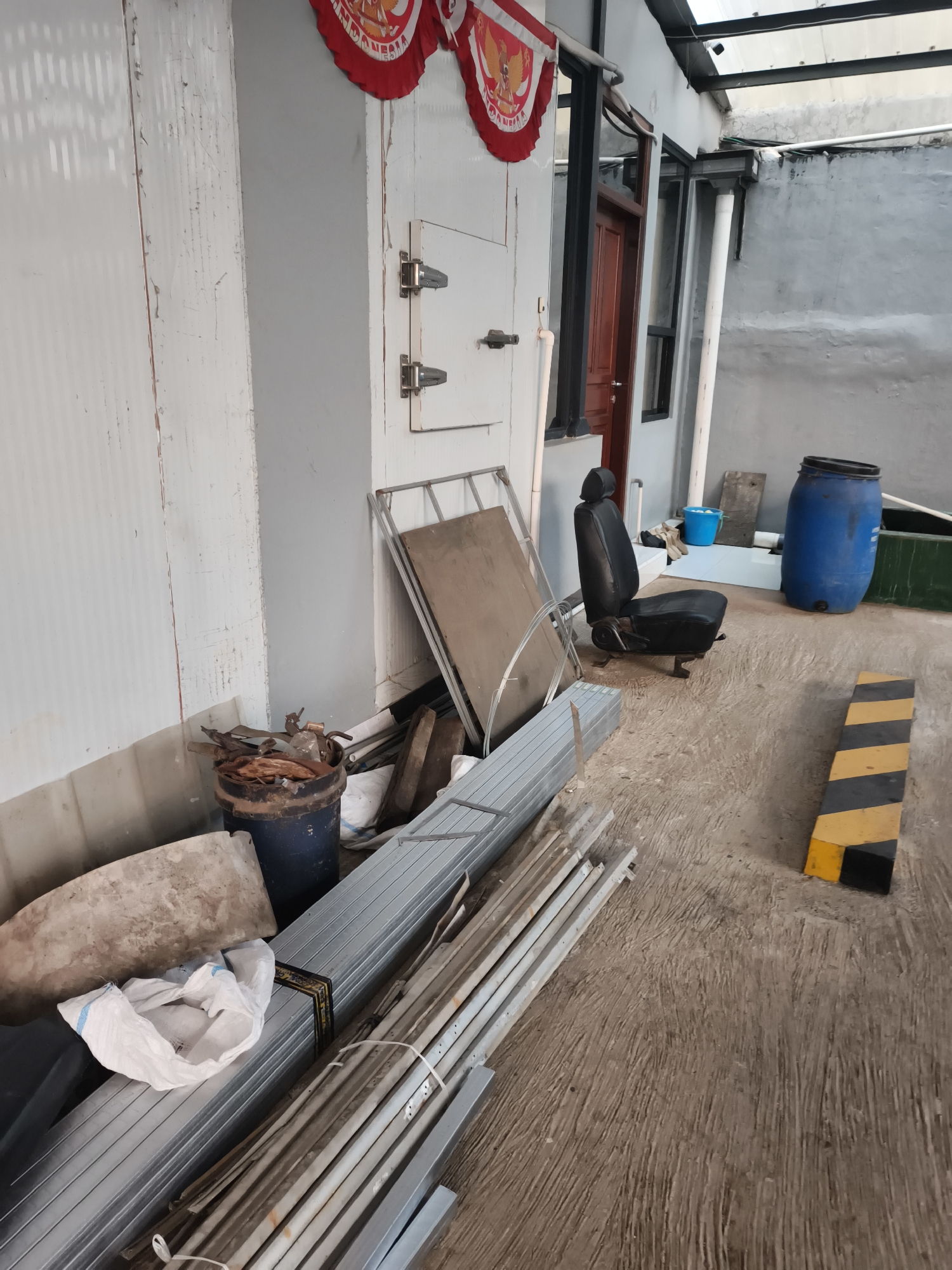The image depicts an outdoor scene, possibly a backyard or alleyway, filled with various construction materials and discarded items. In the foreground, stacks of galvanized metal framing are piled on the concrete ground. To the left, a large, light-colored, irregularly shaped object rests beside a white plastic bag. Further back, a rusty metal drum filled with what appears to be scrap metal and wood is visible. Behind the drum, a black car seat with a headrest is positioned on the ground. To the right of the car seat, a large blue plastic barrel stands upright. In the background, a portion of a building with a grey concrete wall and a corrugated metal roof is present. Red and white decorative banners with what looks like a national emblem are hanging from the building's facade. A yellow and black striped traffic barrier is situated on the right side of the image. The ground is made of textured concrete, showing signs of wear and tear. The overall impression is one of an unorganized storage area or construction site. The location is stated to be in the city of Pekalongan, Indonesia. No people are visible in the image, and no actions or interactions are depicted.