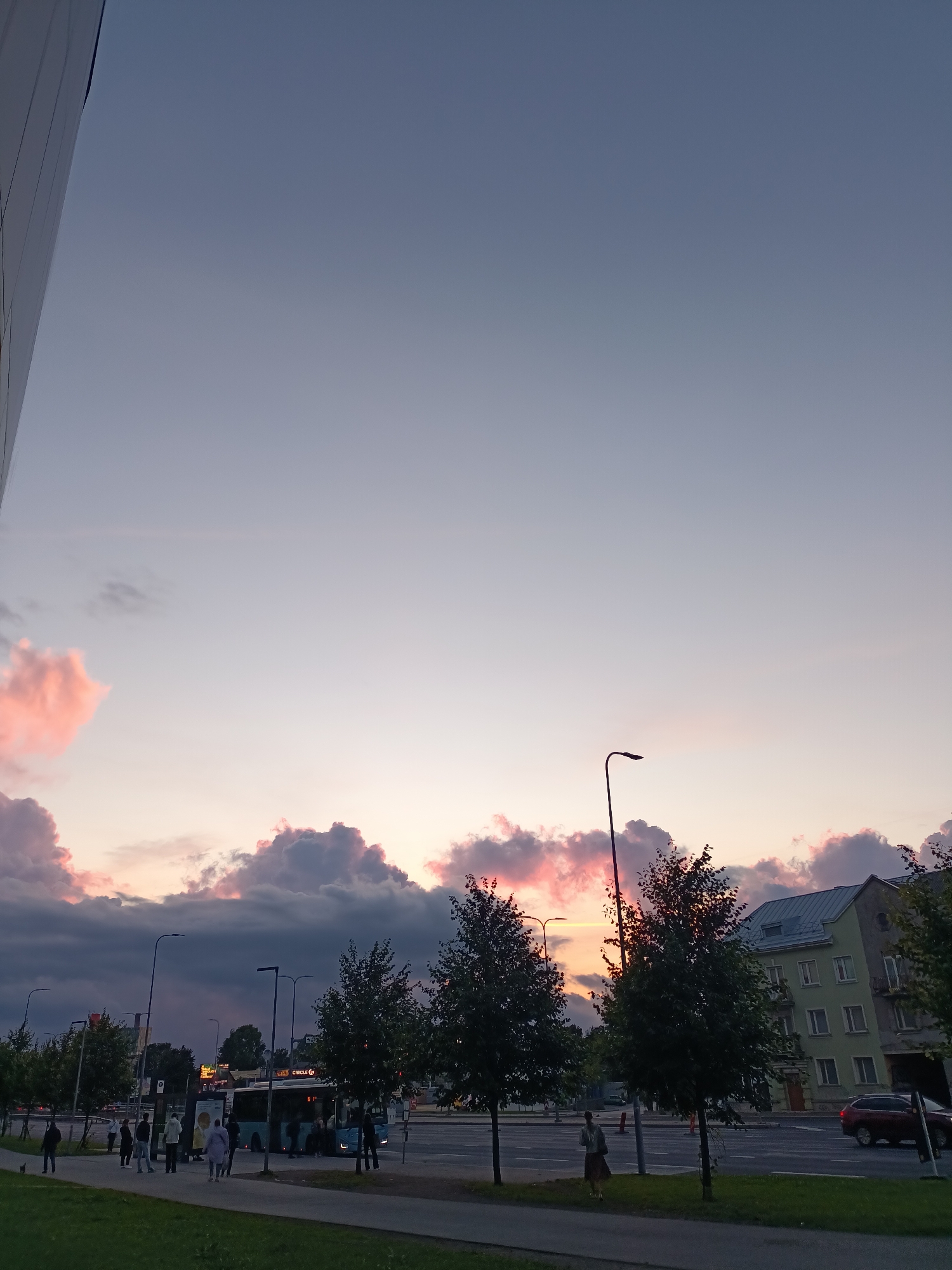The image shows a street scene in Tallinn, Estonia, during sunset. The sky is a gradient of soft purples and pinks, with fluffy clouds tinged with sunset colors. In the foreground, a grassy area and a paved walkway are visible. Further back, a street with cars and a bus is depicted. Several streetlights illuminate the area. People are seen walking along the walkway and waiting at a bus stop. Buildings, including a light green apartment building, line the street. Trees are scattered throughout the scene, some with full foliage and others appearing more sparse. The overall atmosphere is calm and tranquil, characteristic of an evening in a city.