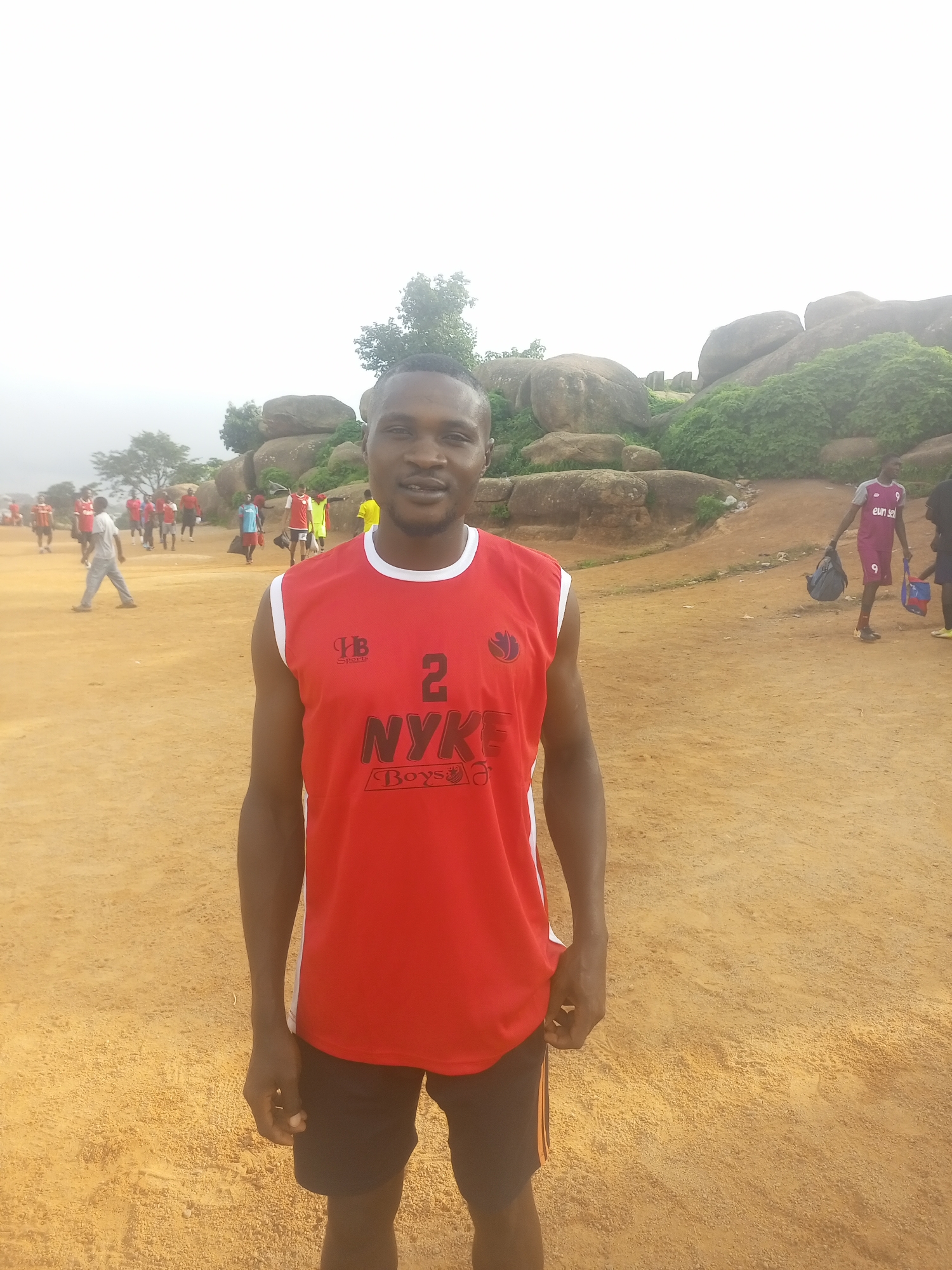 A man stands in the foreground, facing the camera and smiling. He is wearing a red sleeveless jersey with white trim and the number "2" and "NYK Boys" printed on the front, along with a logo. He is also wearing black shorts. The ground is sandy and there are several people in the background, some of whom appear to be playing a sport. In the distance, there are large rock formations covered in greenery. The sky is overcast. The location is the old football field at the Gidan Dare settlement in Jos, Nigeria.