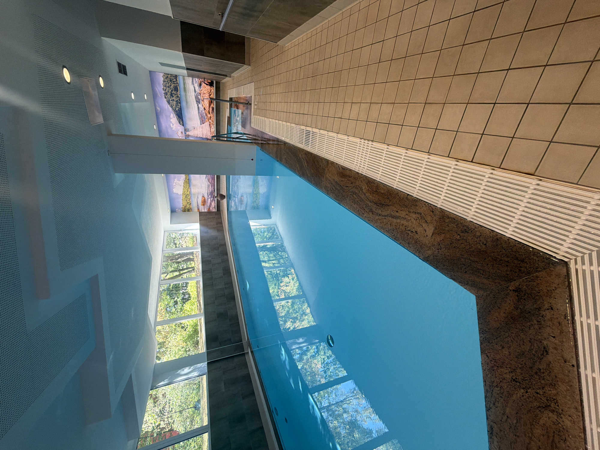 The image shows an indoor swimming pool with a bright blue chlorinated water body. The pool is rectangular and has a dark speckled stone edge. To the left of the pool, there is a large window offering a view of lush green trees and foliage, indicating an outdoor environment. The ceiling of the pool area is a light color with a grid pattern and recessed lighting fixtures. Along the right side of the pool, there is a tiled walkway with a white slatted drainage system bordering the pool. Further to the right, the wall is covered in large, dark tiles. Two murals depicting coastal landscapes with a pier and water are visible on the wall at the far end of the pool area. There are no people visible in the image. This appears to be an indoor swimming pool, possibly located in a hotel or a spa facility.