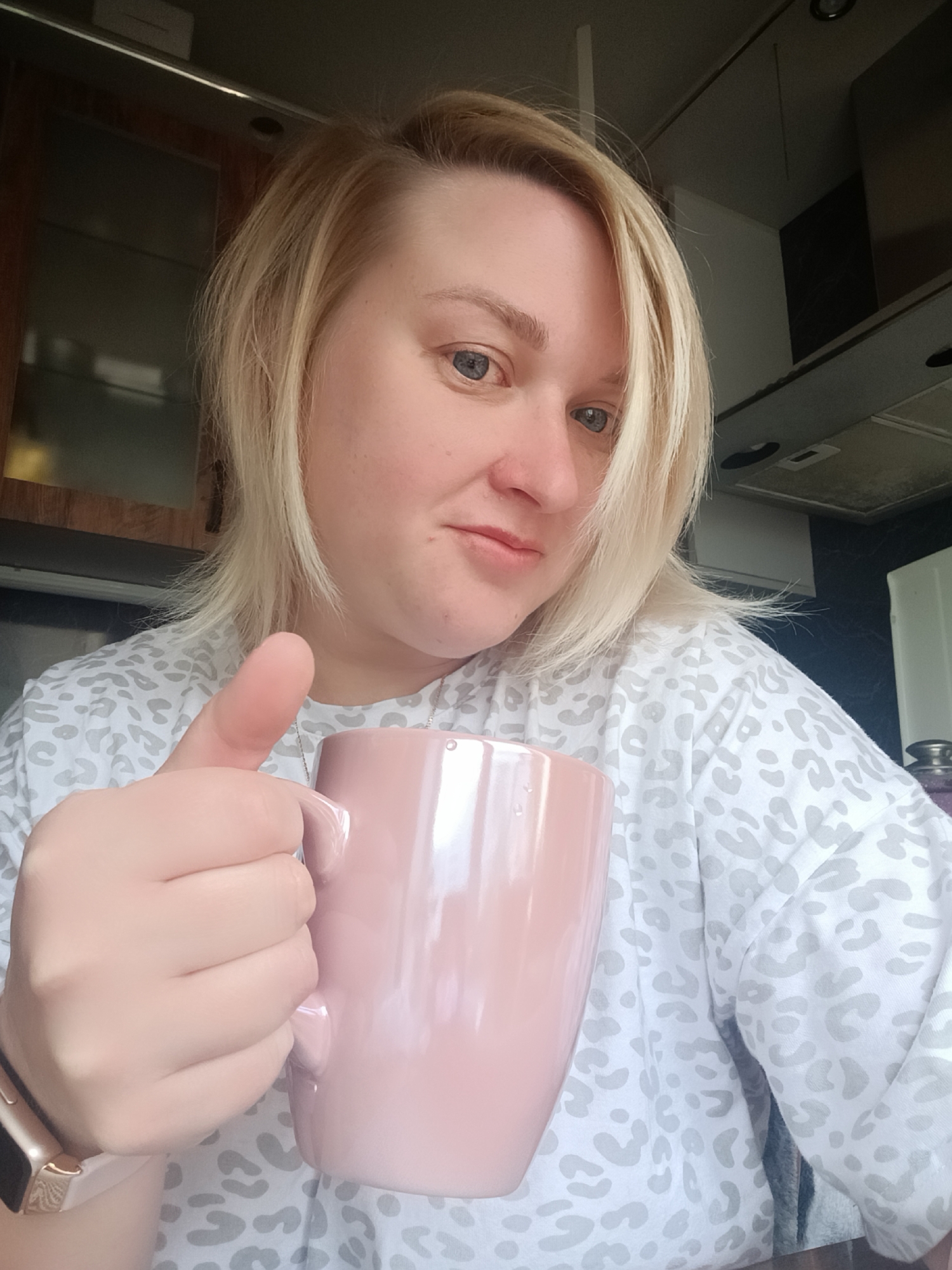 A fair-skinned woman is holding a pink mug, giving a thumbs-up. She has short, blonde hair styled with a side part, and blue eyes. She wears a patterned, long-sleeved shirt and an apple watch. The background shows a kitchen setting with wooden cabinets above and an exhaust fan above. The location appears to be indoors but the image provides no information to indicate the scene's location is in the city of Chaikovsky, Russia.