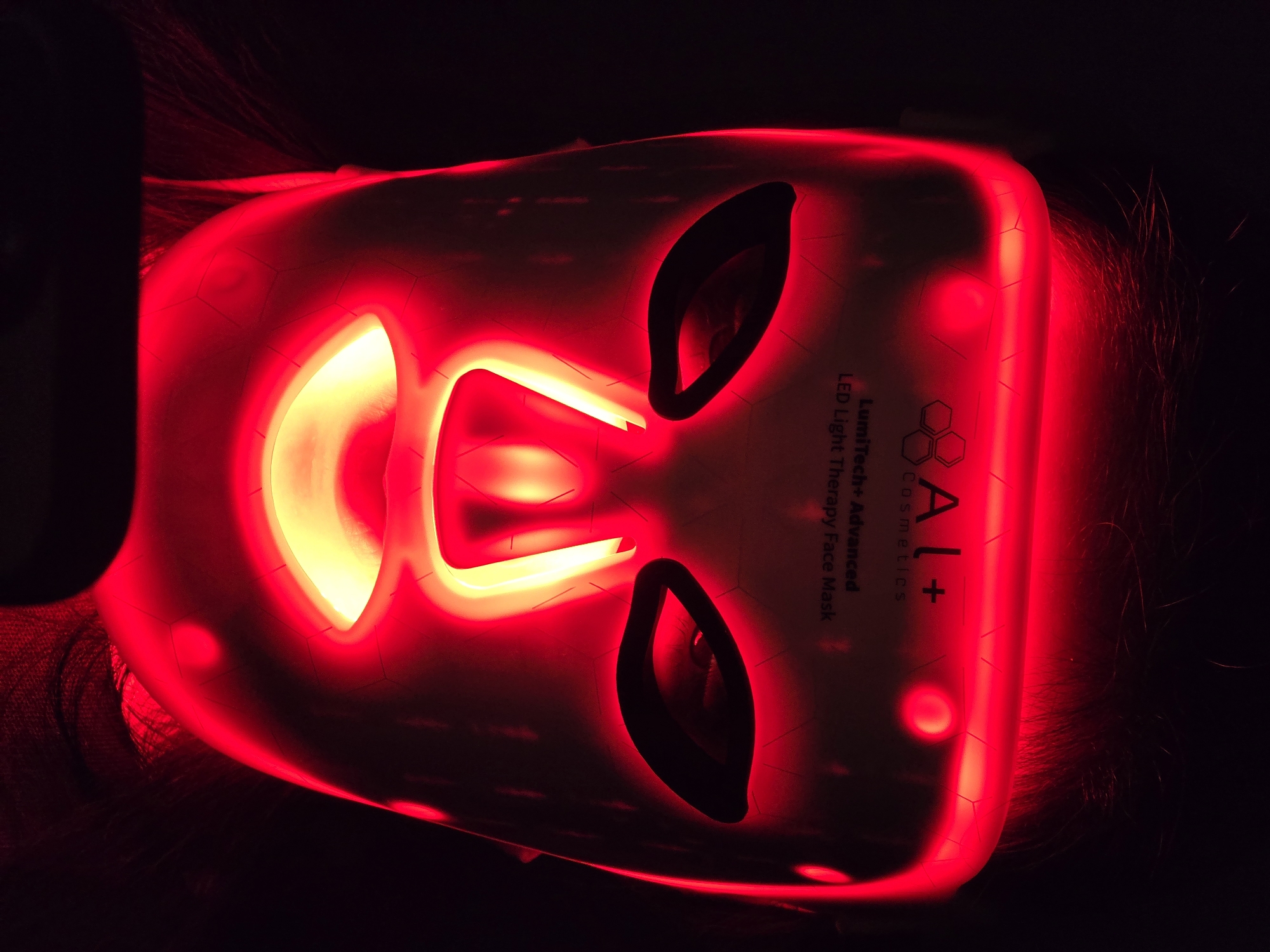 The image presents a close-up, top-down perspective of a person wearing a glowing red LED light therapy face mask in a dark environment. The mask covers the wearer's face, with dark hair visible around its perimeter. The primary illumination from the mask is a vibrant red light, which becomes a brighter, yellowish-orange hue around the bridge of the nose and mouth area. Dark, almond-shaped openings serve as eye holes, revealing no discernible facial features beneath.
The surface of the mask features a subtle hexagonal grid pattern. Text is visible on the right side of the mask, oriented vertically, stating "LumiTech+ Advanced LED Light Therapy Face Mask" and "BAL+ Cosmetics," accompanied by a logo composed of two intertwined honeycomb-like shapes. The intense glow against the deep black background suggests the treatment is taking place in a dimly lit room, likely at night. The activity depicted is a personal beauty or skincare treatment utilizing LED light technology. The scene is situated in Plovdiv, Bulgaria.