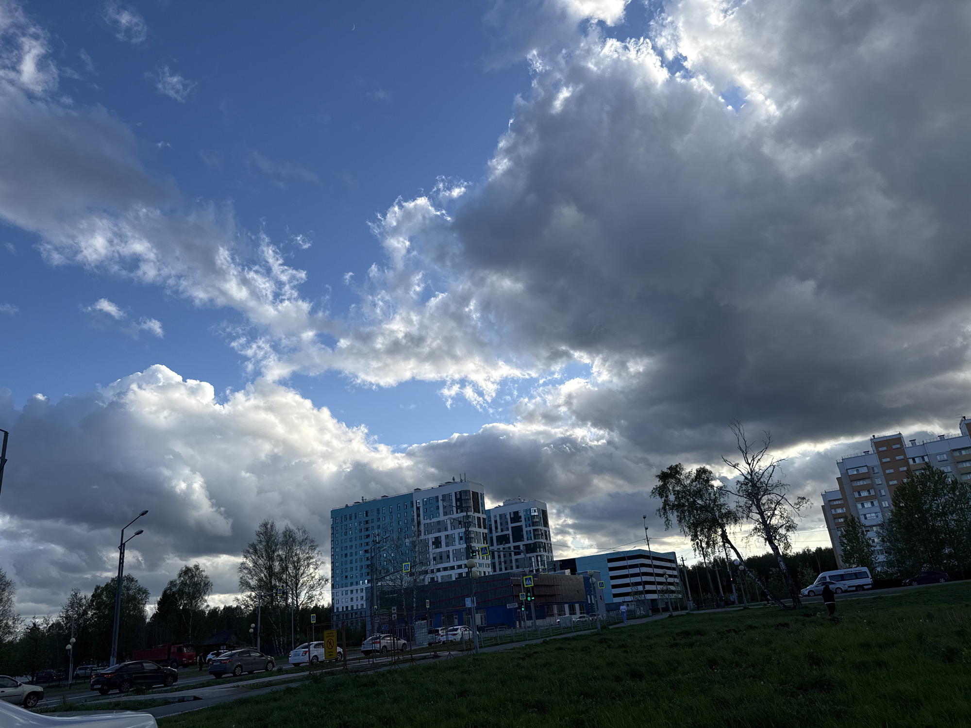 The image depicts a cloudy sky over a cityscape in Tobolsk, Russia. Cumulus and cumulonimbus clouds dominate the sky, with patches of blue visible. Below, buildings are visible, including a modern blue and white high-rise apartment complex and a lower-rise brick building. A street with several parked and moving vehicles runs in front of the buildings, and there are some green trees visible in the foreground and middle ground. A grassy area is in the immediate foreground. Streetlights are present. The overall lighting suggests late afternoon or early evening.