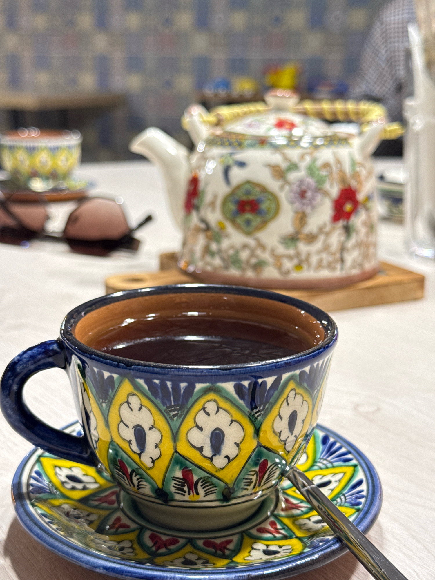 A close-up shot focuses on a vibrantly decorated teacup and saucer filled with dark liquid, likely tea. The cup and saucer feature intricate patterns of blue, yellow, green, and red, with floral motifs. A silver spoon rests on the saucer. In the background, a white teapot with similar decorative elements sits on a wooden coaster. Out-of-focus sunglasses are visible to the left, and a blurred background suggests an indoor setting with patterned wallpaper and furniture. The image does not contain information about the city of Chaikovsky, Russia.