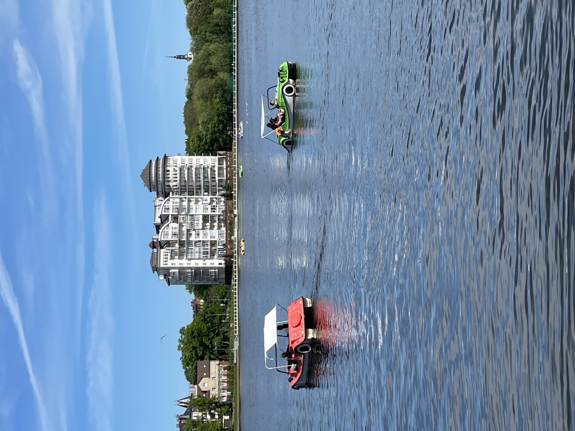 The image shows a cityscape on the shore of a body of water, likely in Kaliningrad, Russia, as suggested by the architectural style of the buildings and the presence of recreational pedal boats.
On the water, there are two prominent pedal boats. One is red with a white awning, and another is green with white tires. Occupants are visible on both boats.
The shoreline features a mix of buildings, including residential houses and a large, multi-story building with a distinct, white façade. Trees and other greenery line the shore behind the buildings. In the far distance, a church spire or tower is visible, adding to the city's skyline.
The sky is clear and blue, with light clouds creating contrast. The water has a textured surface reflecting the ambient light and surrounding objects.