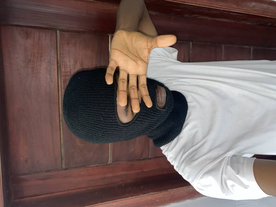 The image displays a single individual in Joma, Ghana, positioned against a background of reddish-brown wooden paneling. The individual is wearing a black knitted balaclava that covers the entire head and face, with openings for the eyes and mouth. A plain white, short-sleeved t-shirt is visible. The person's right hand is raised, palm outwards, with fingers slightly spread, partially obscuring the eye openings of the balaclava. The visible skin on the hand and through the balaclava openings is dark-toned. The wooden background shows visible grain and appears to be composed of vertical planks with horizontal joints.