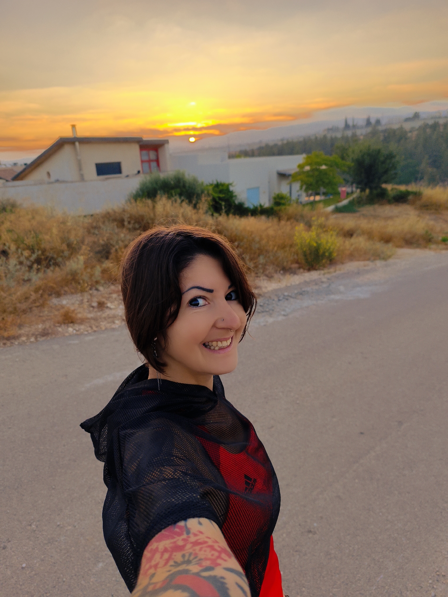 A woman with short dark hair and a nose ring smiles at the camera, her face partially visible. She is wearing a black mesh top over a red athletic shirt, and her left arm, adorned with tattoos, is extended towards the viewer. The woman stands on an asphalt road with dry vegetation on the left and a few buildings in the background. The scene is illuminated by the warm glow of a sunset over a hilly landscape, suggesting a rural or semi-rural setting within the Lachish Regional Council, Israel. The sky is a vibrant mix of orange and yellow hues, with the sun visible near the horizon.