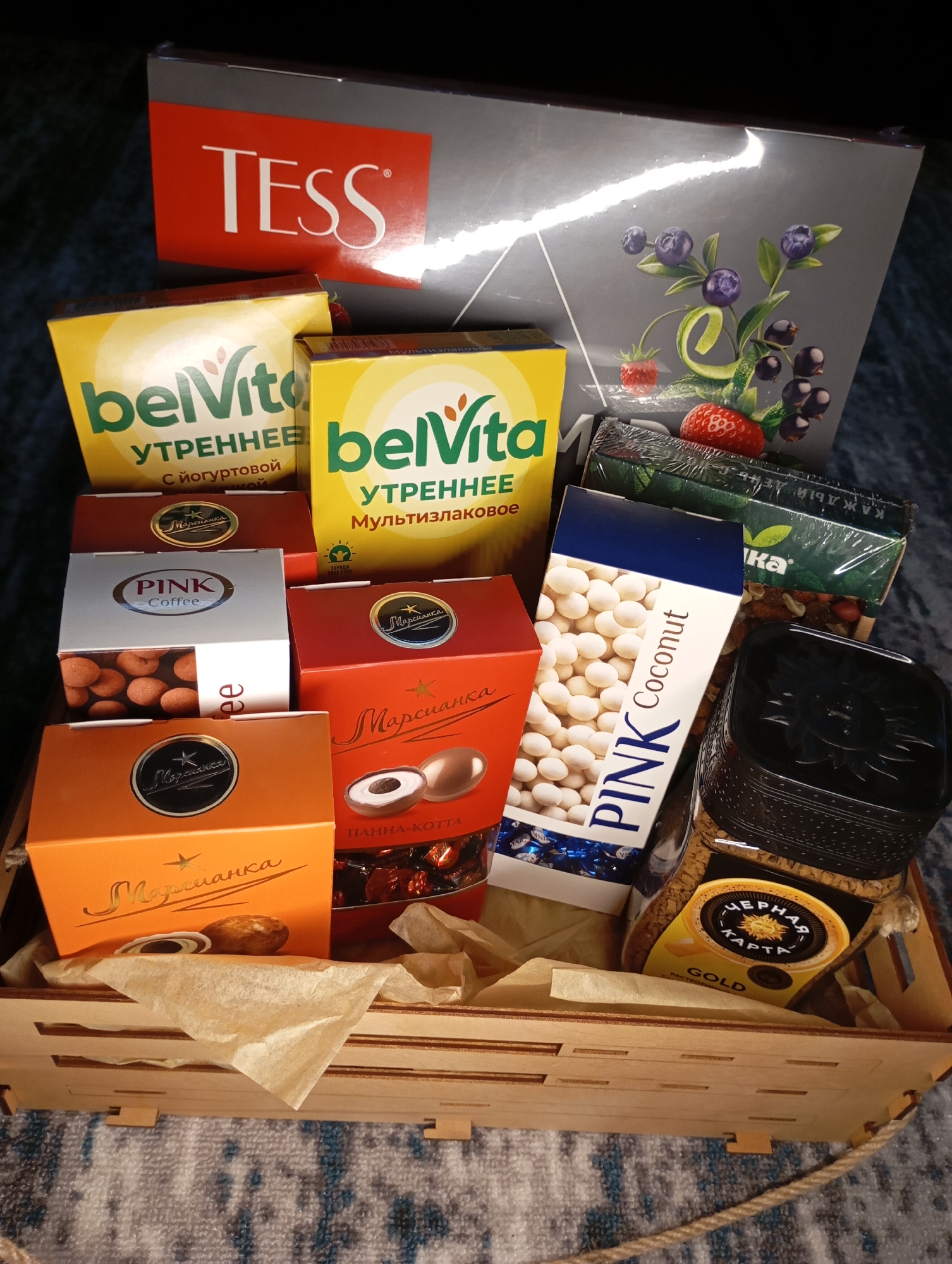 A wooden crate filled with assorted food products. The crate contains two boxes of "belVita" brand breakfast biscuits, one "TESS" brand tea box, three boxes of "Марсианка" brand candies, one "PINK Coffee" brand candy box, one "PINK Coconut" brand candy box, and one jar of "ЧЕРНАЯ КАРТА" brand instant coffee. The crate is lined with crumpled beige tissue paper. The background is a dark, textured surface.