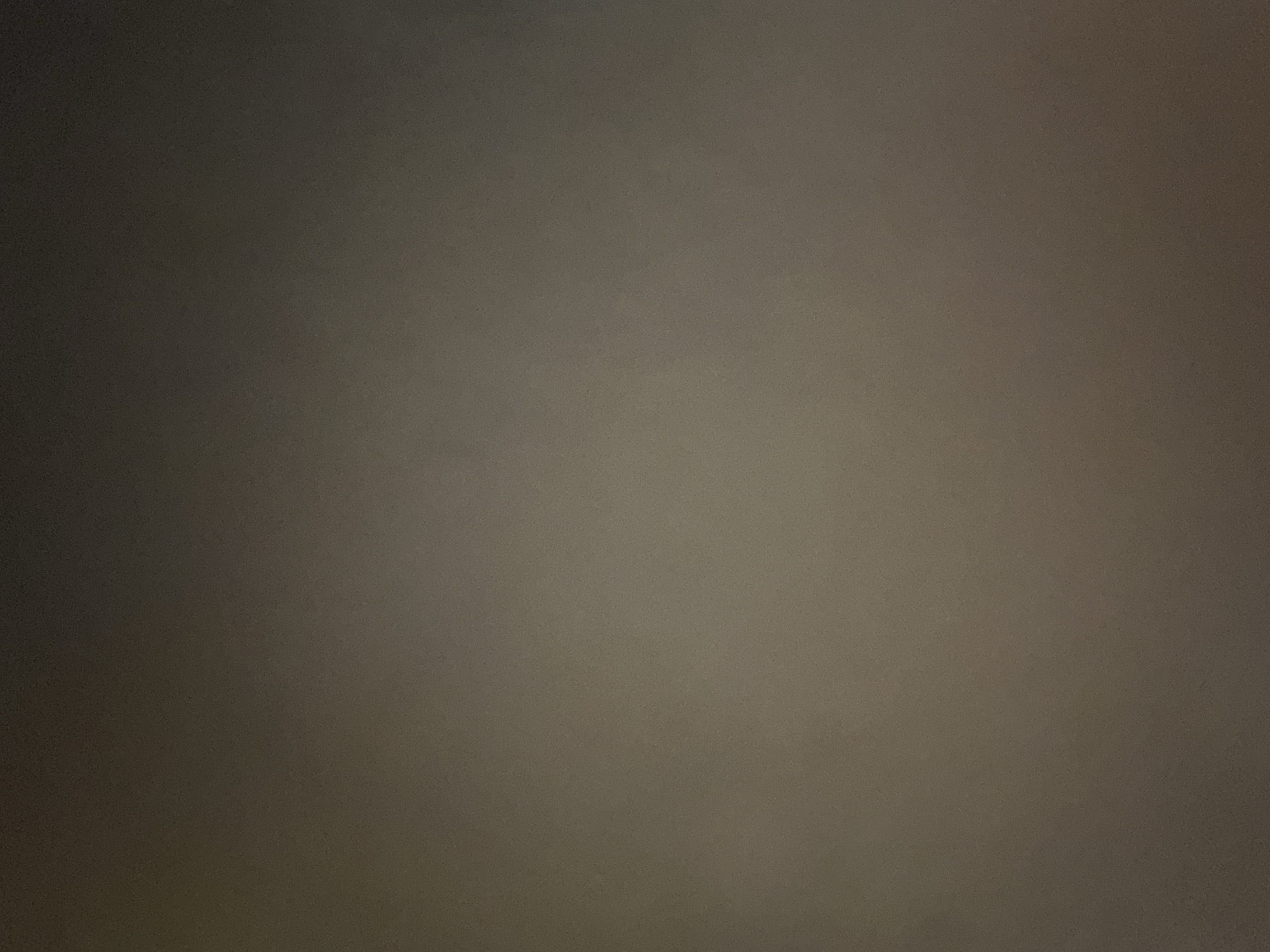 This is a still image of a plain, textured surface. The color of the surface is a muted, brownish-grey, with subtle variations in shade creating a gradient. The left side of the image is darker, appearing to be in shadow, while the right side is slightly lighter and brighter. The texture appears grainy, suggesting it might be fabric, painted wall, or a similar material. There are no discernible subjects, objects, or a specific scene depicted. No activity or event is taking place. There is no visible text, and no clear cues about the time of day, weather, or specific location are provided. The overall impression is of a neutral, abstract background.