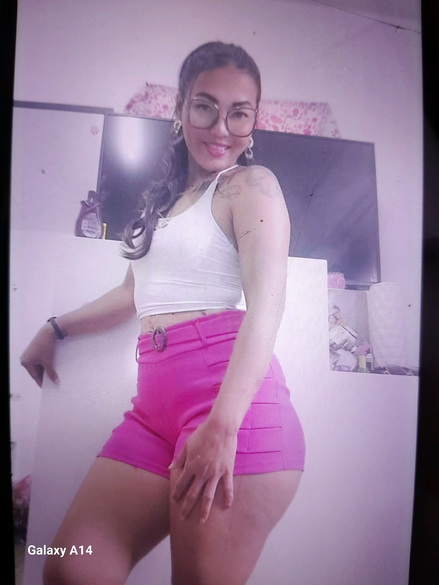 A young woman with dark hair and glasses poses for a photo indoors. She is wearing a white crop top and bright pink shorts. Her left arm is bent at the elbow and rests on a white surface, while her right hand is placed on her right thigh. The background includes a television, a shelf with various items, and a pink patterned fabric. The image is watermarked with "Galaxy A14". The location is identified as Noord, Suriname.