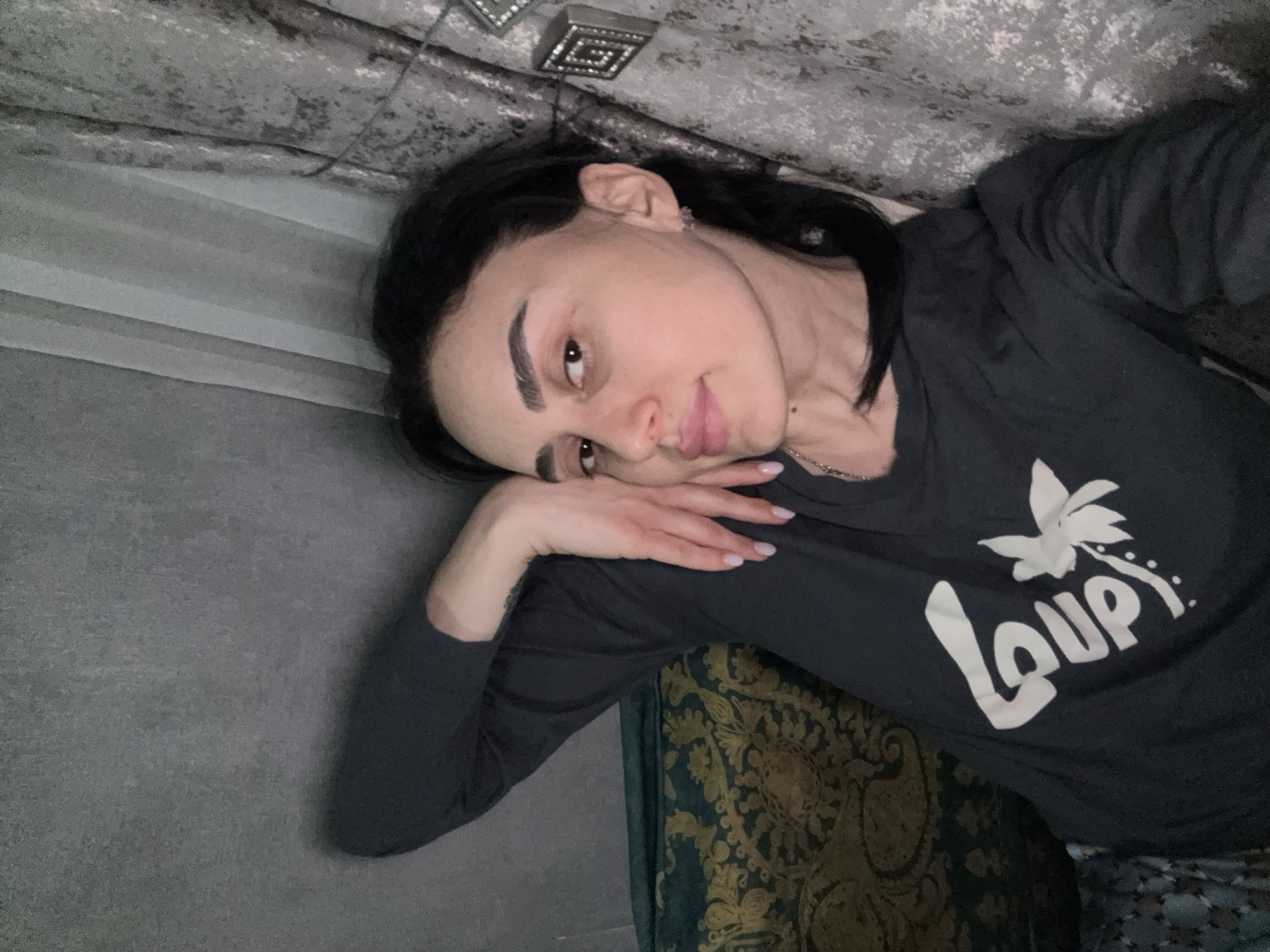 A woman with dark hair and fair skin is lying down, resting her head on her hand. She is wearing a dark long-sleeved shirt with a white graphic of the word "Loup." She is lying on a patterned fabric, possibly a bedspread or curtain. The background includes a gray wall, a decorative ceiling fixture, and a textured ceiling. The location is not specified as being in Tbilisi, Georgia, and therefore cannot be confirmed.