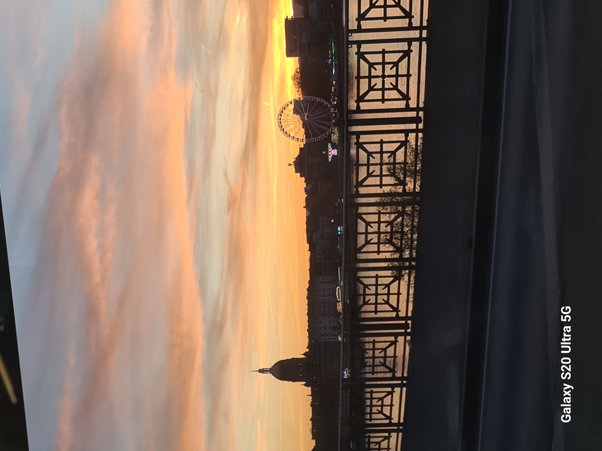 The image depicts a sunset in Wiesbaden, Germany, with the city skyline and the Nerobergbahn ferris wheel visible against an orange sky. A decorative metal railing is in the foreground, likely from a bridge. The Schloßplatz dome is also visible in the background.