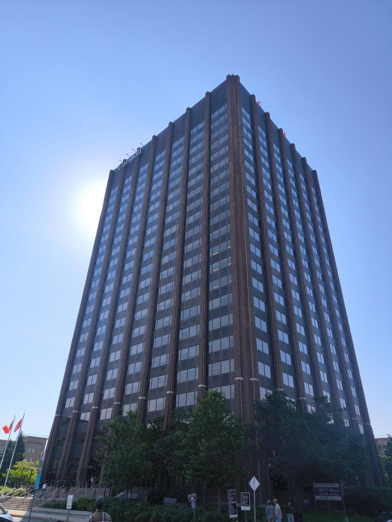 The image shows the exterior of a tall, dark-colored office building with a repeating pattern of windows. The building appears to be the "Canada Post Building" located at 390 Slater Street in Ottawa, Canada. The text "Canada" is visible in large letters on the top of the building. The sun is shining brightly from behind the building, creating lens flare. Red flags are visible at the top of the building, and Canadian flags are positioned near the entrance. Trees and greenery are present at the base of the building, and a street and sidewalk are visible in the foreground. A few people are seen near the entrance and on the sidewalk. The sky is clear and blue.