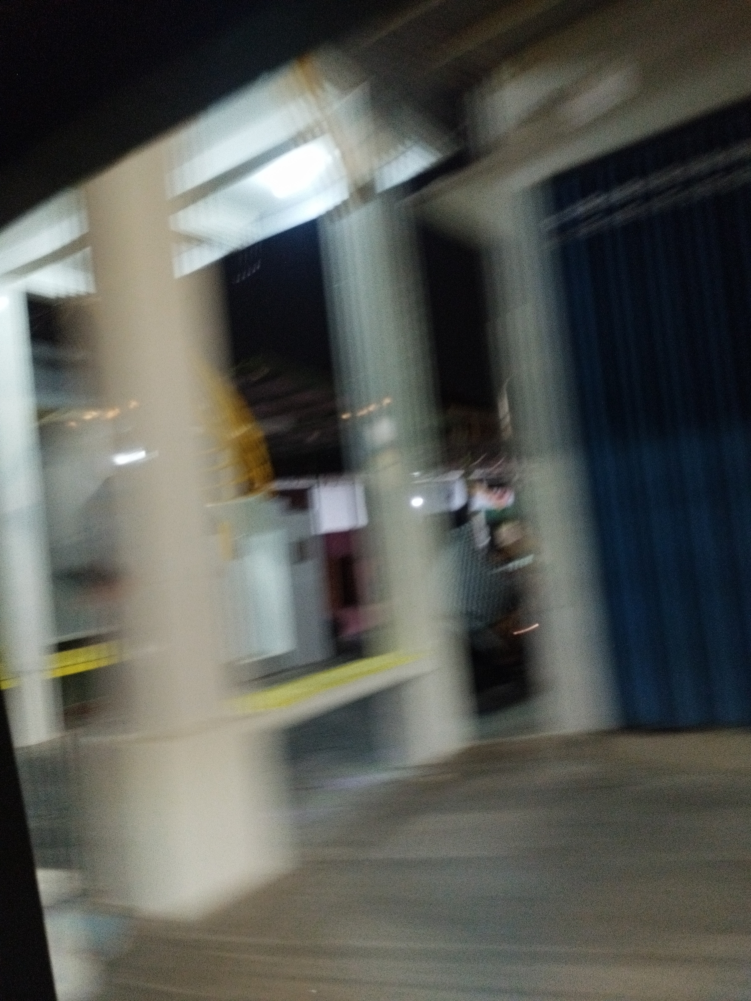 The image appears to be taken from inside a vehicle in motion, looking out at an outdoor, covered area at night. The motion blur is significant, making specific details difficult to discern. In the foreground, a blurry, light-colored pillar is visible on the left. A bright yellow line runs horizontally across the scene, possibly marking a boundary or safety line.
Further into the scene, there are structures that suggest a commercial or industrial setting. A large, dark blue, corrugated metal door or wall is prominent on the right side of the image. Behind the pillar and to the left, there are glimpses of what might be buildings or shelters with white roofs and some internal lighting. The lighting is dim and artificial, contributing to the overall dark ambiance.
Based on the context provided, the location is specified as the city of Surabaya, Indonesia. The scene could depict an area like a loading dock, a warehouse entrance, or a covered walkway in a commercial or industrial zone within Surabaya. The time of day is clearly night due to the darkness and artificial lighting. There are no clearly identifiable people or specific actions taking place due to the extreme motion blur and low light conditions.