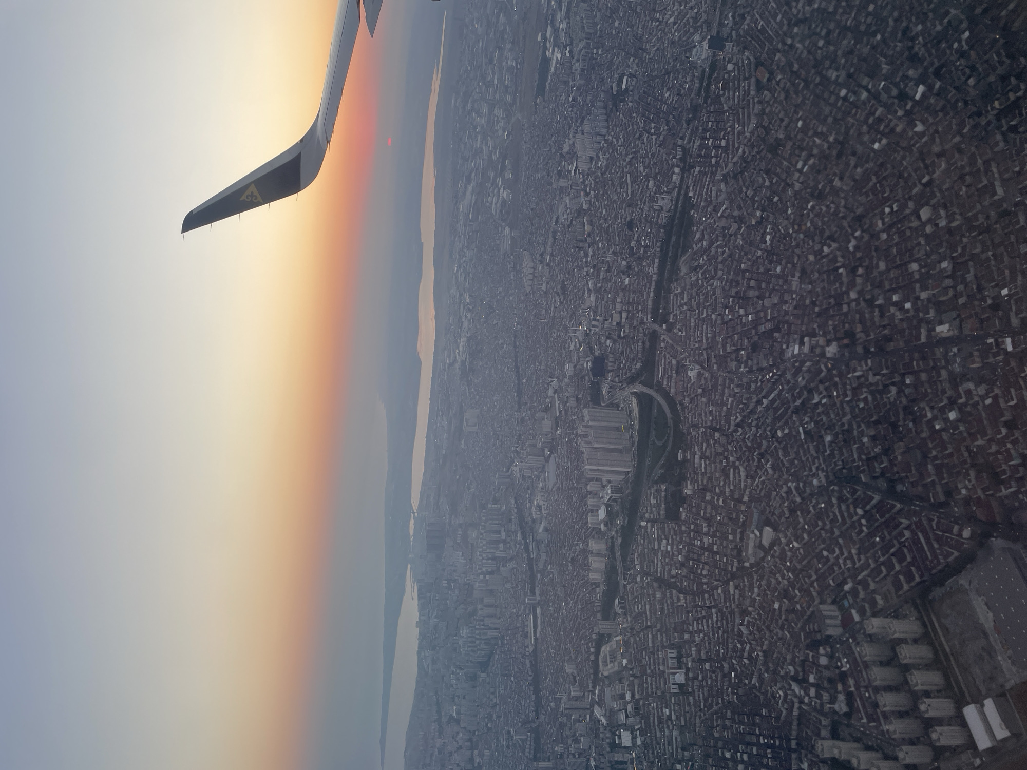 An aerial view shows a dense urban landscape with a body of water in the distance, viewed from an airplane window. The sky exhibits a gradient of colors from light blue at the top to soft orange and pink hues near the horizon, indicating either sunrise or sunset. The wing of an airplane, featuring a dark blue tip with a yellow emblem, is visible in the upper right portion of the frame, partially obscuring the sky. The city below is characterized by a vast expanse of tightly packed buildings with dark roofs, interspersed with some taller, modern structures. A river or canal winds through the city. The scene captures a sprawling metropolis at dusk or dawn, with the atmospheric light casting a serene glow over the urban sprawl. This image is likely of Beykoz, Turkey, given the geographical features and urban density often associated with the area.