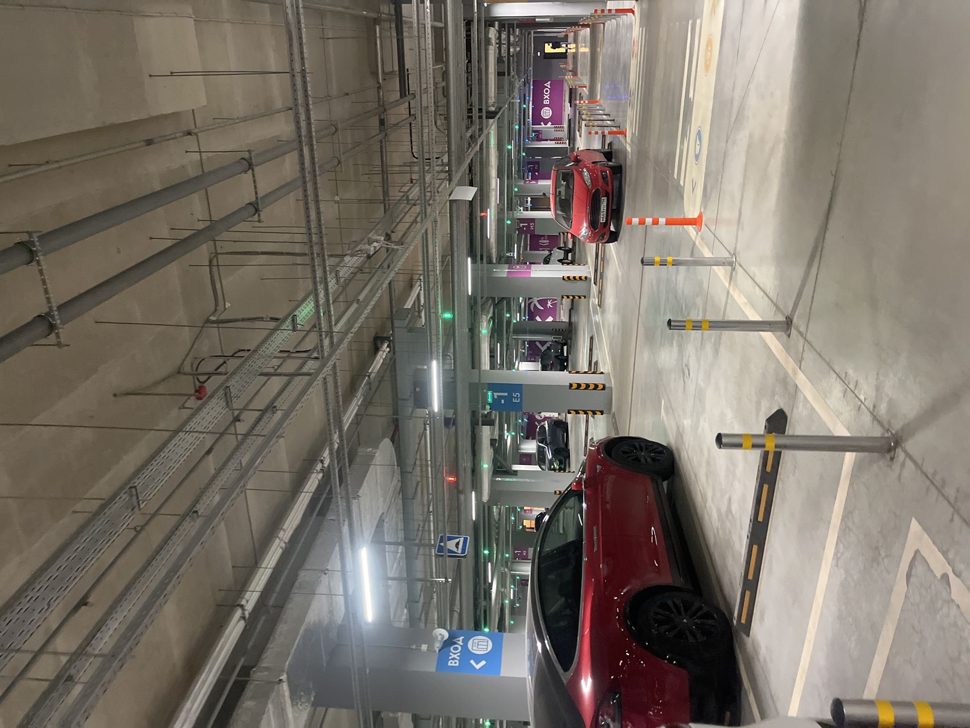 The image depicts the interior of a parking garage in Saint Petersburg, Russia, illuminated by fluorescent lights. The garage ceiling is exposed, showing a network of pipes and electrical conduits. Two red vehicles are prominently positioned. One is parked to the right, while the other is located further down the aisle. Various signs and informational markers are visible along the walls, some with the Russian word "ВХОД" (entrance). Physical barriers and traffic cones are used to delineate parking spaces and pathways.