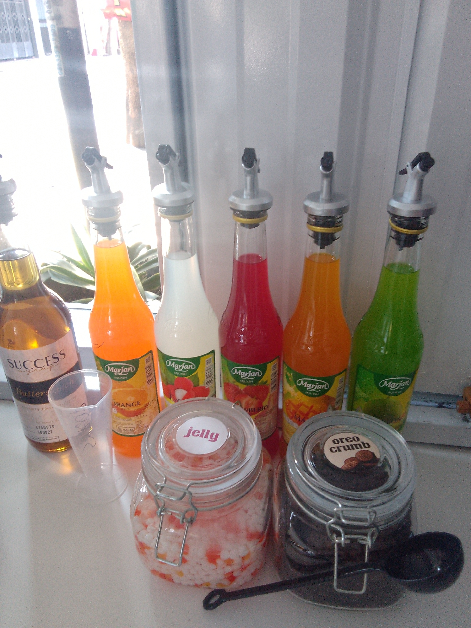 In Trenggalek, Indonesia, on a white countertop, a collection of beverage syrups and jars is displayed. Six tall, narrow bottles with silver pour spouts are arranged in a row. From left to right, the first bottle contains amber liquid, labeled "SUCCESS Buttery flavour." The second bottle is filled with bright orange liquid and is labeled "Marjan Orange Squash." The third bottle holds white liquid and is labeled "Marjan Squash." The fourth bottle contains vibrant red liquid and is labeled "Marjan Strawberry Squash." The fifth bottle has yellow-orange liquid and is labeled "Marjan Mango Squash." The sixth bottle is filled with lime green liquid and is labeled "Marjan Squash." To the left of the bottles, a clear plastic measuring cup is positioned. In front of the bottles, two glass jars with clip-top lids are placed. The left jar contains a pink and white granular substance, with a circular label reading "jelly" in pink text. The right jar is filled with dark brown powder, featuring a circular label with the text "oreo crumb" and an image of chocolate cookies. A black plastic spoon rests on the countertop beside the right jar. The background consists of a white textured surface, possibly a wall or curtain, with a window allowing natural light to illuminate the scene from the left. Behind the bottles, a green plant with long leaves is visible through the glass.