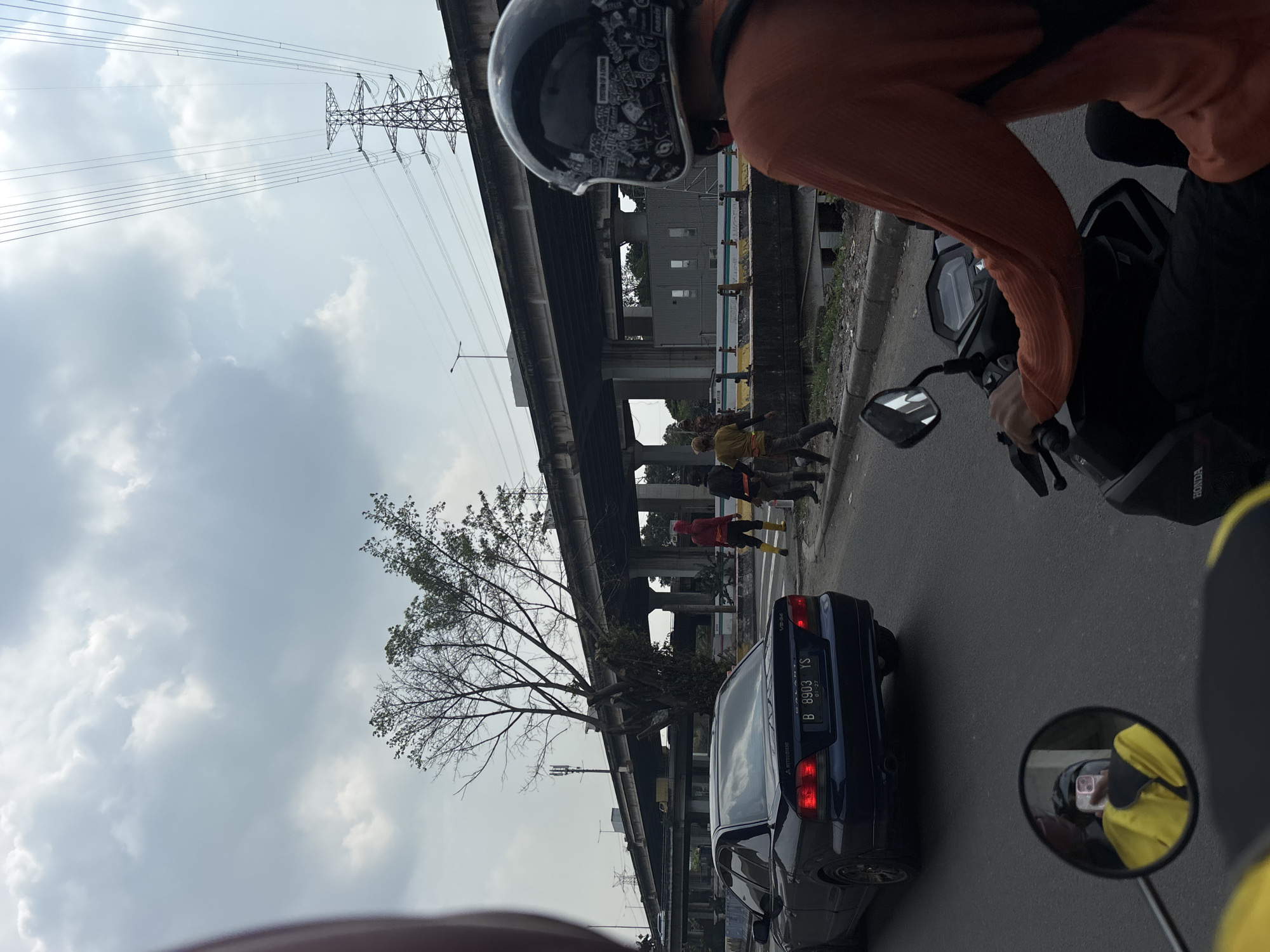 The image, taken from the perspective of a motorcyclist in motion, captures a scene along a road in North Jakarta, Indonesia. The focus is on the traffic and surrounding infrastructure.
A dark blue Mitsubishi Galant with license plate B 8903 YS is visible, positioned to the left of the motorcyclist. Above the road is a concrete overpass, and utility lines extend above the overpass, connecting to a distant power transmission tower.
Several people are visible on the side of the road beyond the overpass. They are wearing various colored clothing.
The sky is overcast with thick, grey clouds, suggesting a potentially rainy day. A motorcycle helmet with stickers is prominent in the foreground, and a side mirror reflects the yellow helmet of another motorcyclist behind the photographer.