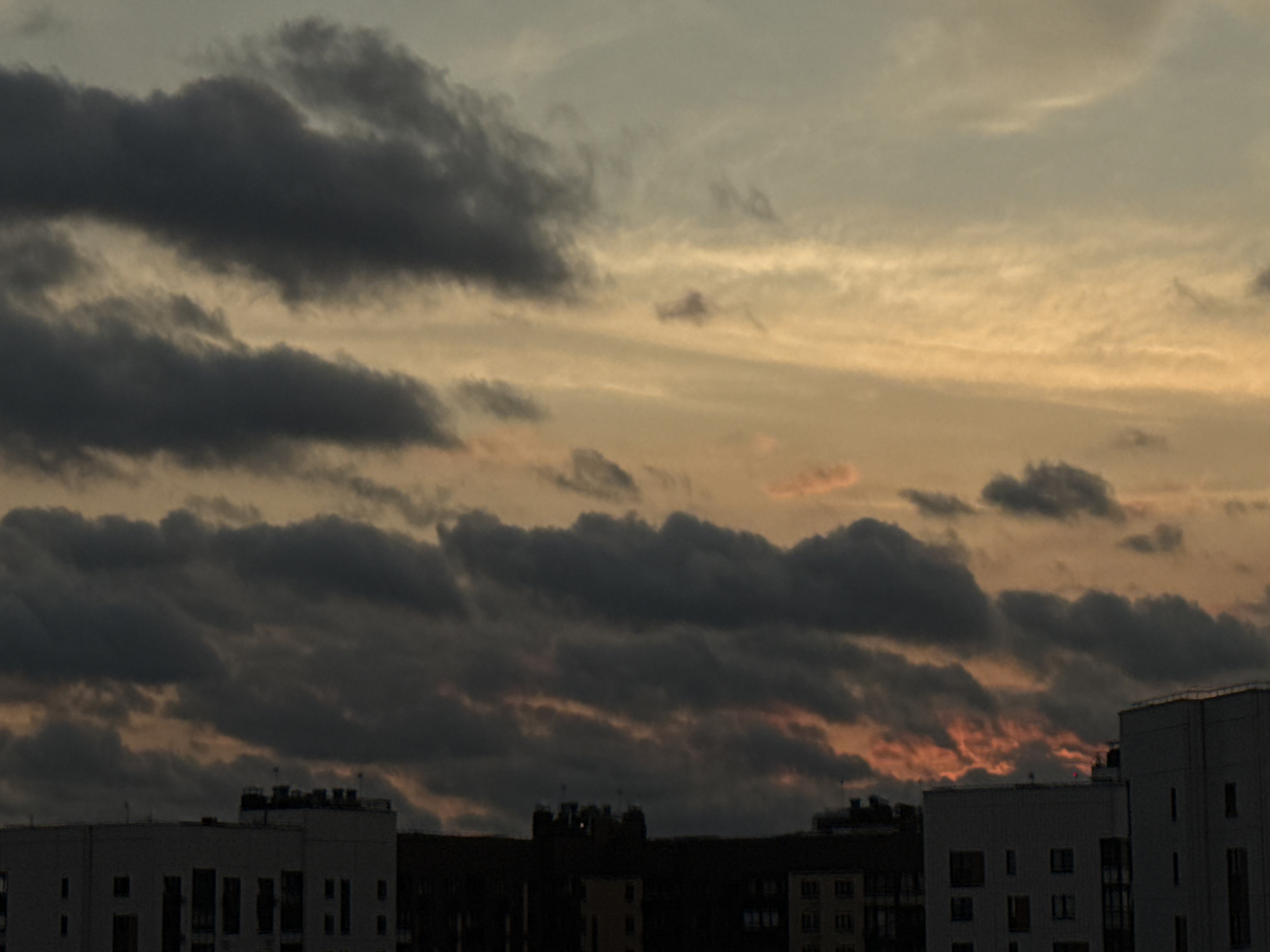 The image captures a view of buildings and a dramatic sky over Lyubertsy, Russia. The sky is filled with a mix of dark, heavy clouds and lighter, wispy formations illuminated by an orange and yellow sunset. The clouds appear to be thick and possibly indicative of approaching weather. In the foreground, the tops of several modern residential buildings are visible. These buildings are mostly white and have multiple stories with numerous windows. Some of the buildings have visible rooftop structures, such as vents or chimneys. The overall scene suggests an urban environment at dusk, with the natural light of the sunset contrasting with the man-made structures. There are no people or specific actions depicted in the image.