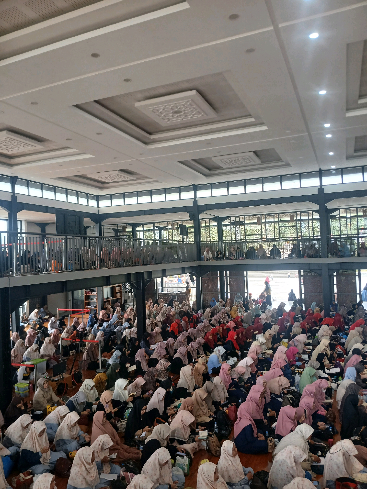 In Arjasari, Indonesia, this image depicts a large gathering of people, predominantly women, seated on the floor of what appears to be a spacious indoor venue, possibly a mosque or community hall. The individuals are wearing hijabs and modest clothing in various colors, with a concentration of pink, white, and beige. Many are looking down, some with books or notes in front of them, suggesting a lesson, prayer, or religious service is taking place. The room has a high ceiling with modern lighting fixtures and decorative paneling. A second level or balcony is visible, also populated by people. The overall atmosphere appears to be one of focused attention and community participation.
